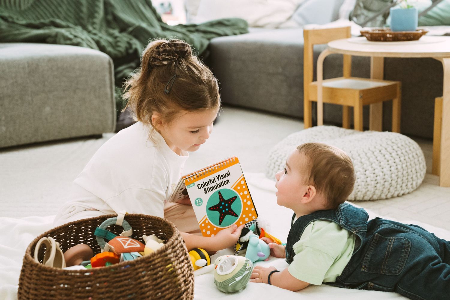 Two children lying on the floor looking at a picture book together next to a basket of toys.