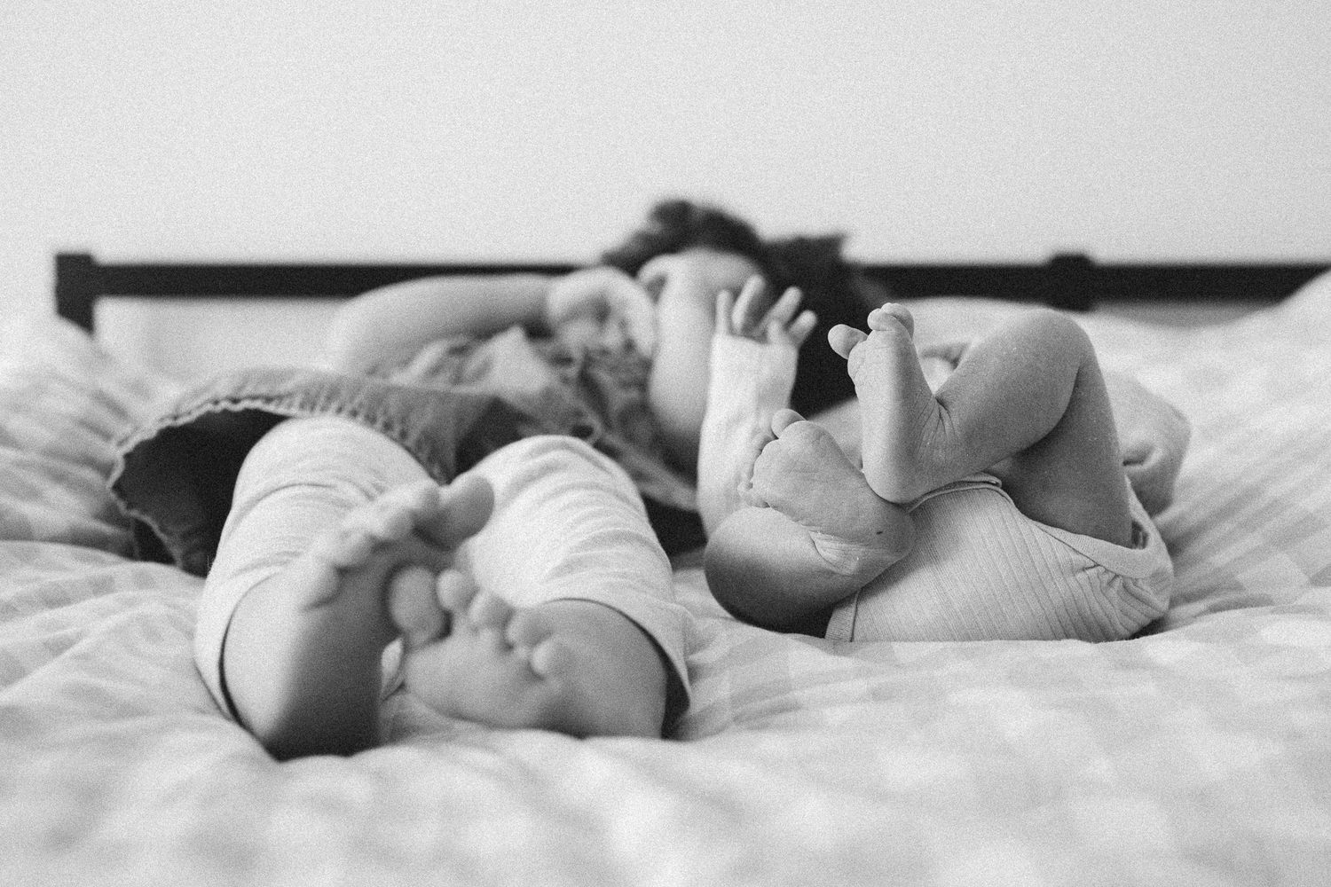 Black and white photo of siblings lying on bed with newborn baby feet in foreground.