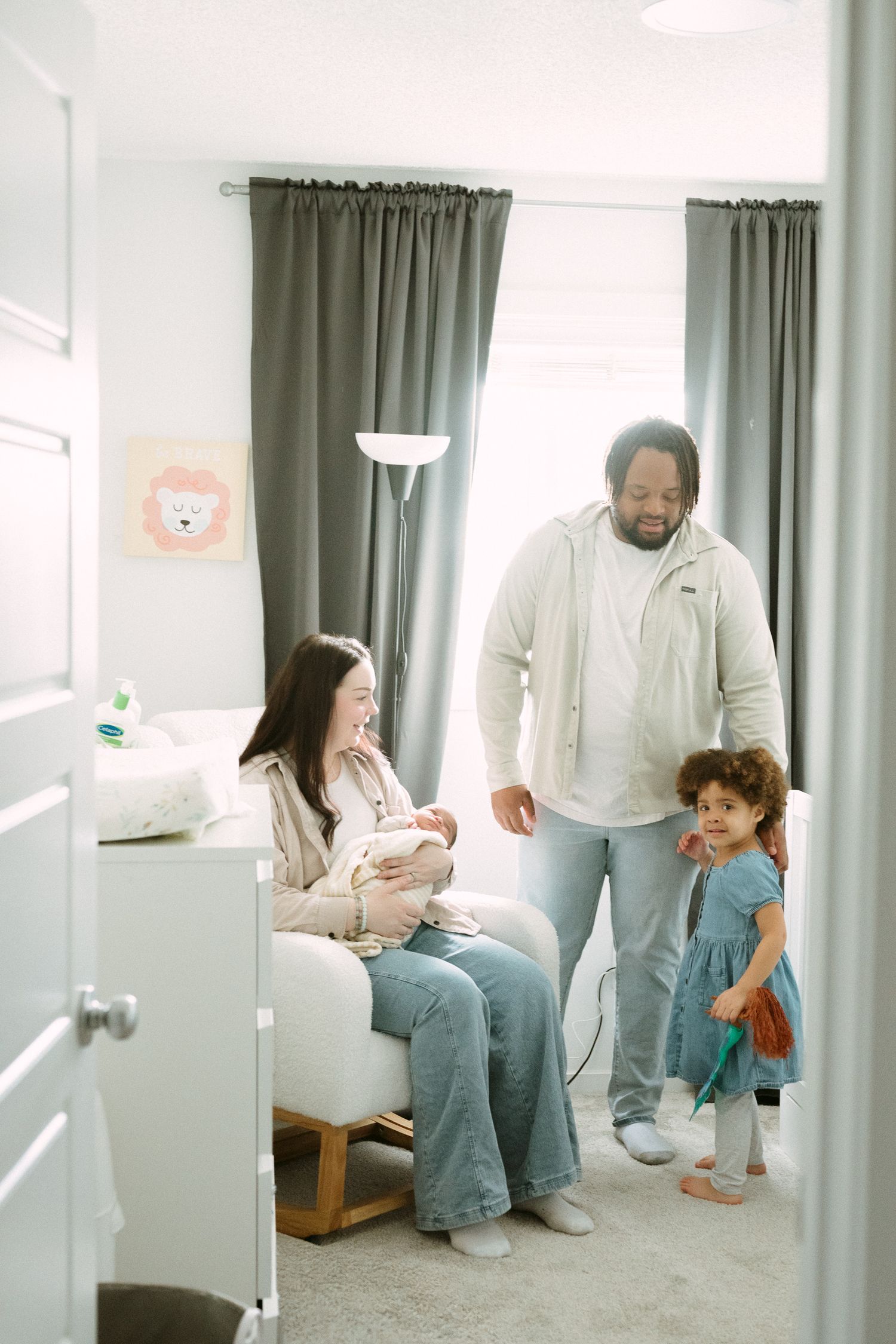 A happy interracial family with a toddler and newborn baby gather in a softly lit nursery room.
