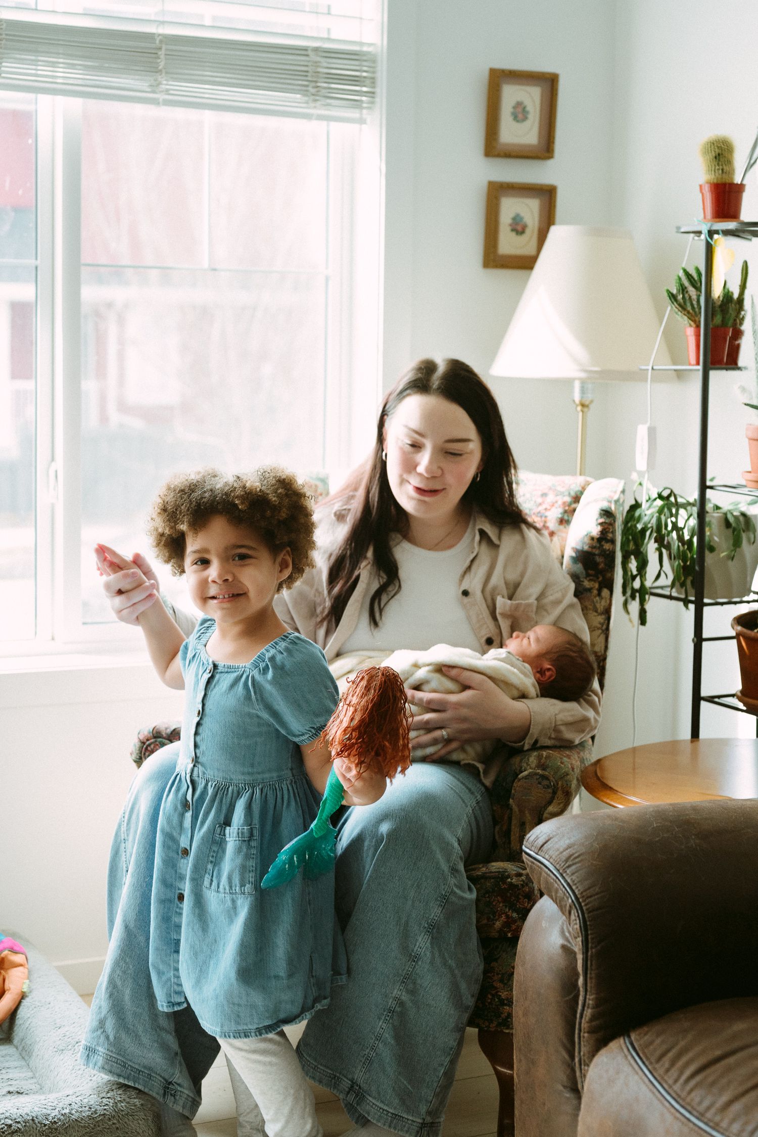 A woman sits in an armchair holding a newborn while a young girl in a blue dress stands beside her in a bright living room.