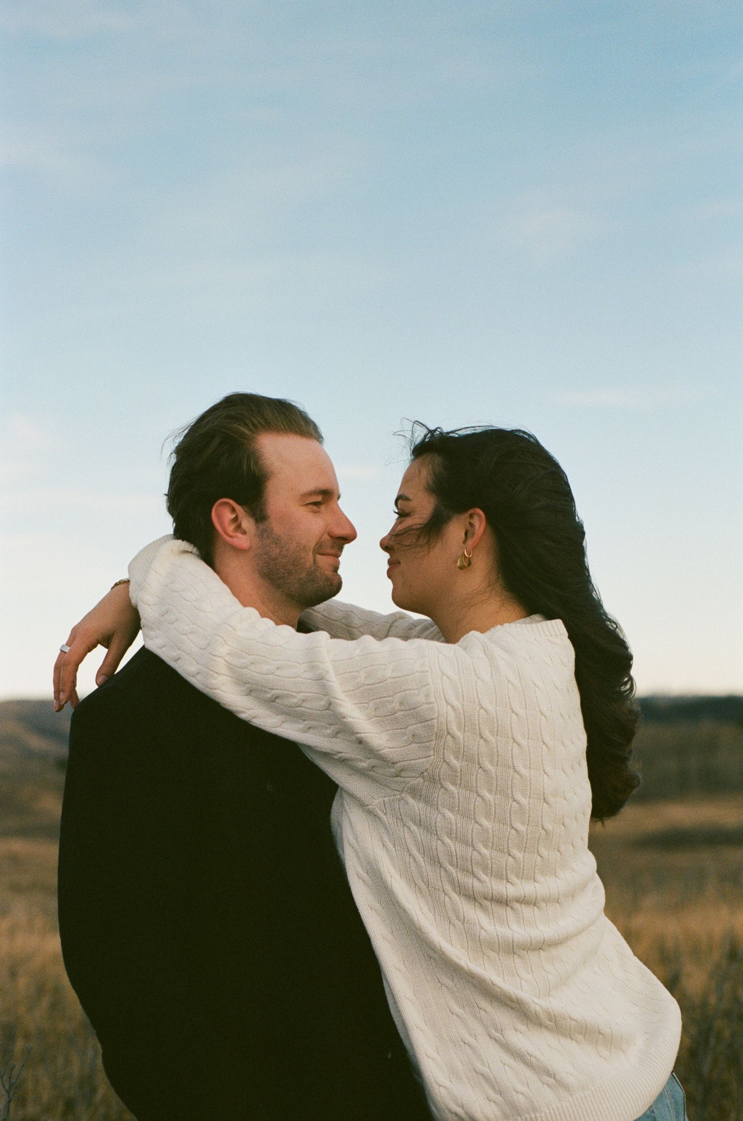 A romantic couple embraces in a field during sunset with the person in white leaning in for a kiss.