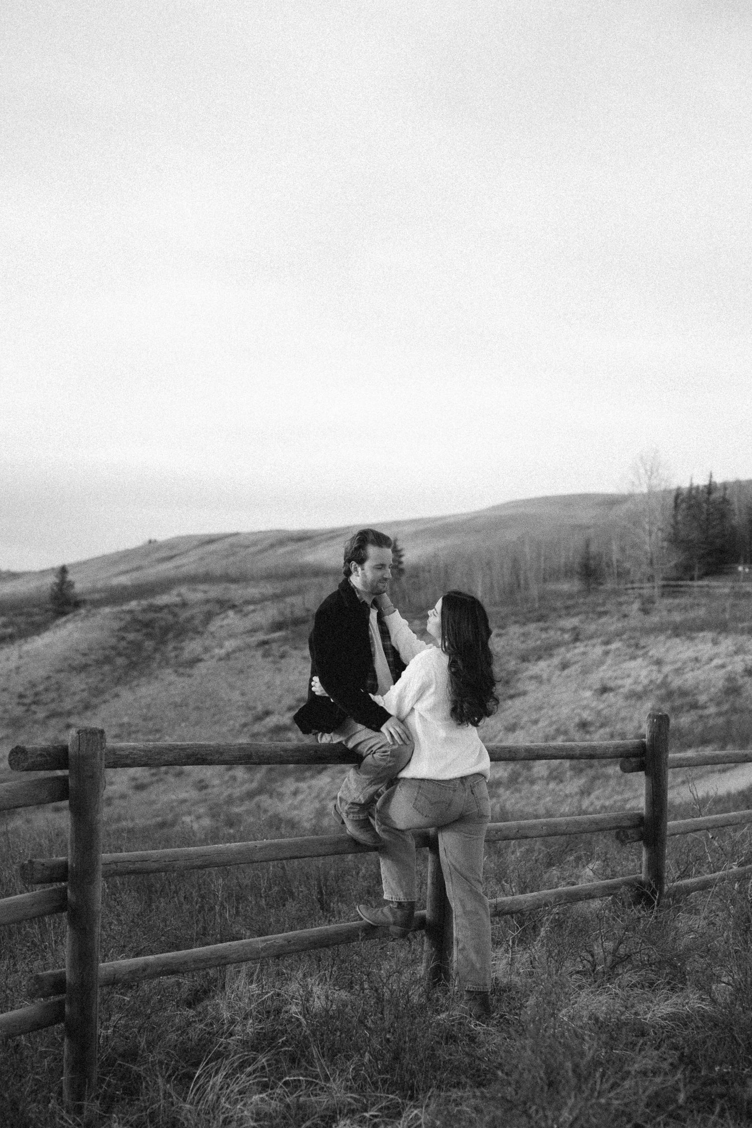 A romantic black and white photo of a couple sitting on a wooden fence in a rural hillside setting.