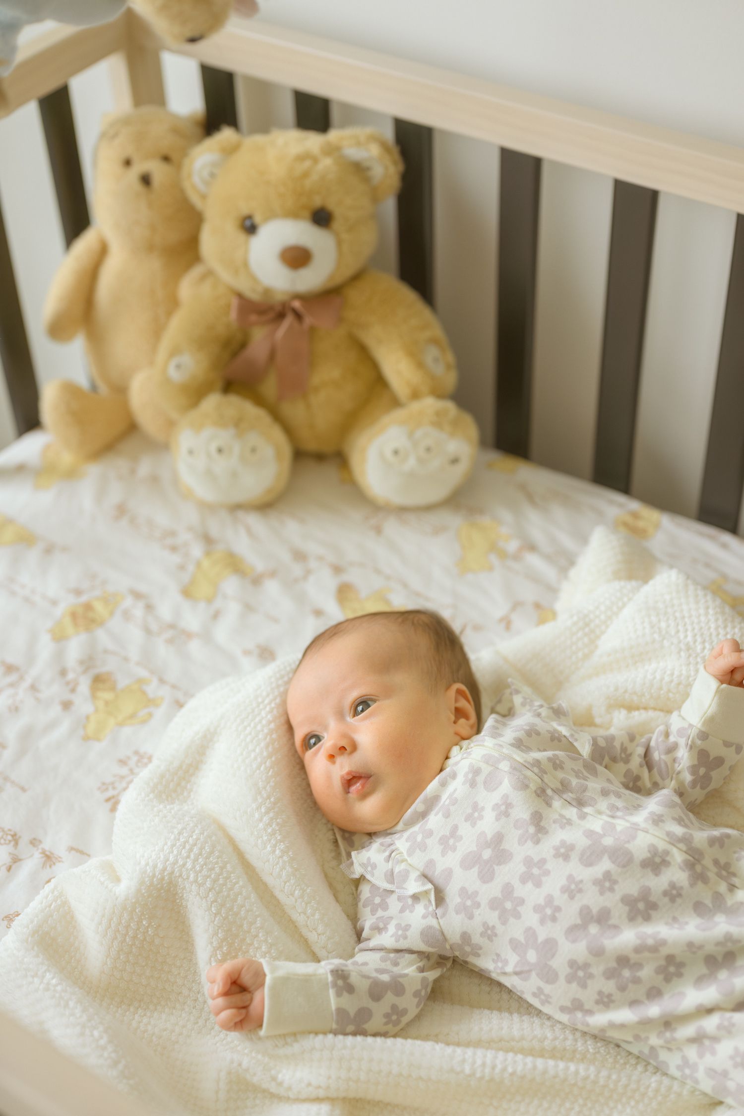 Baby lying in crib with teddy bears and white knit blanket.