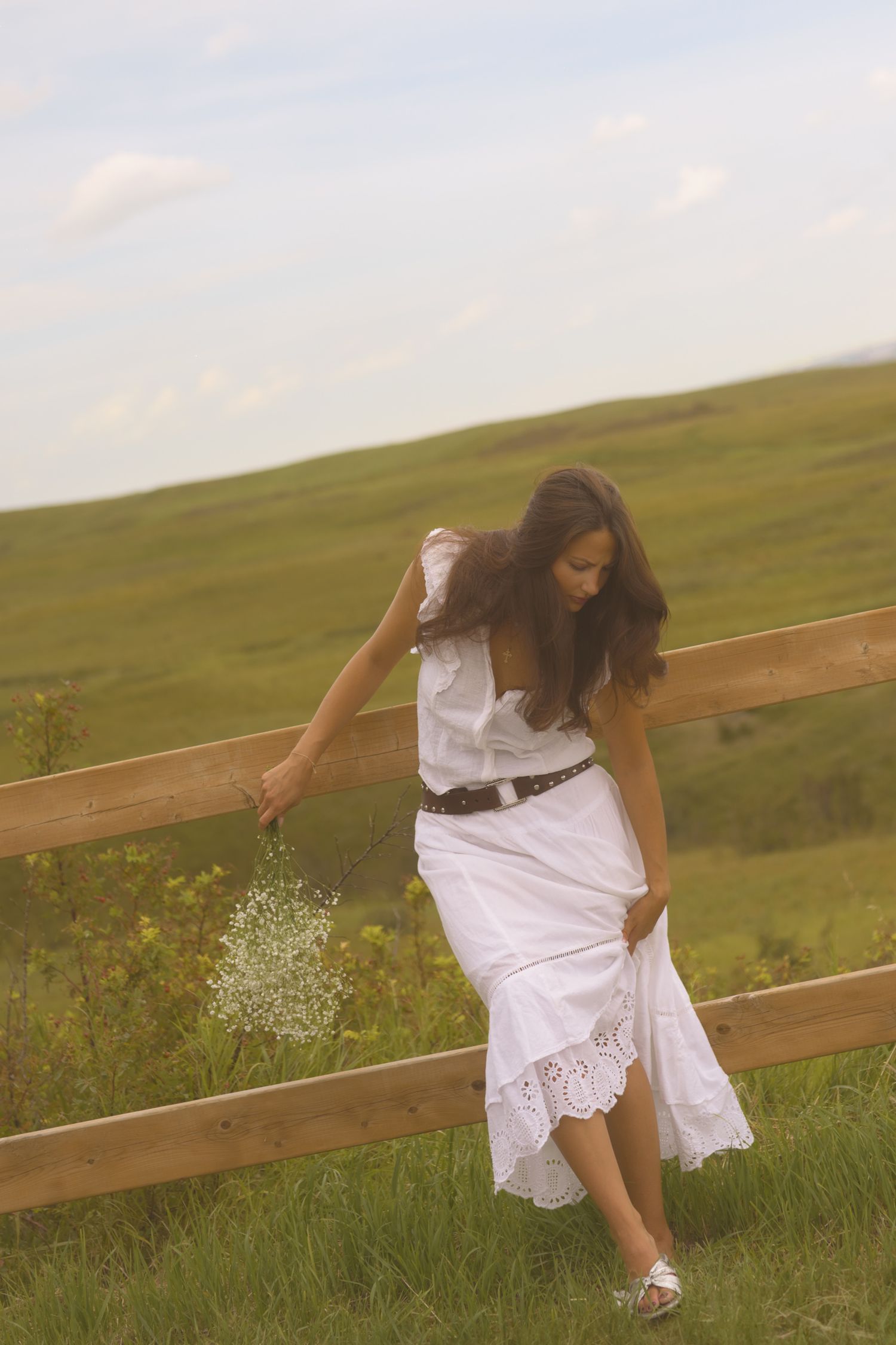 Person in white dress leans on wooden fence surrounded by wildflowers and rolling hills.