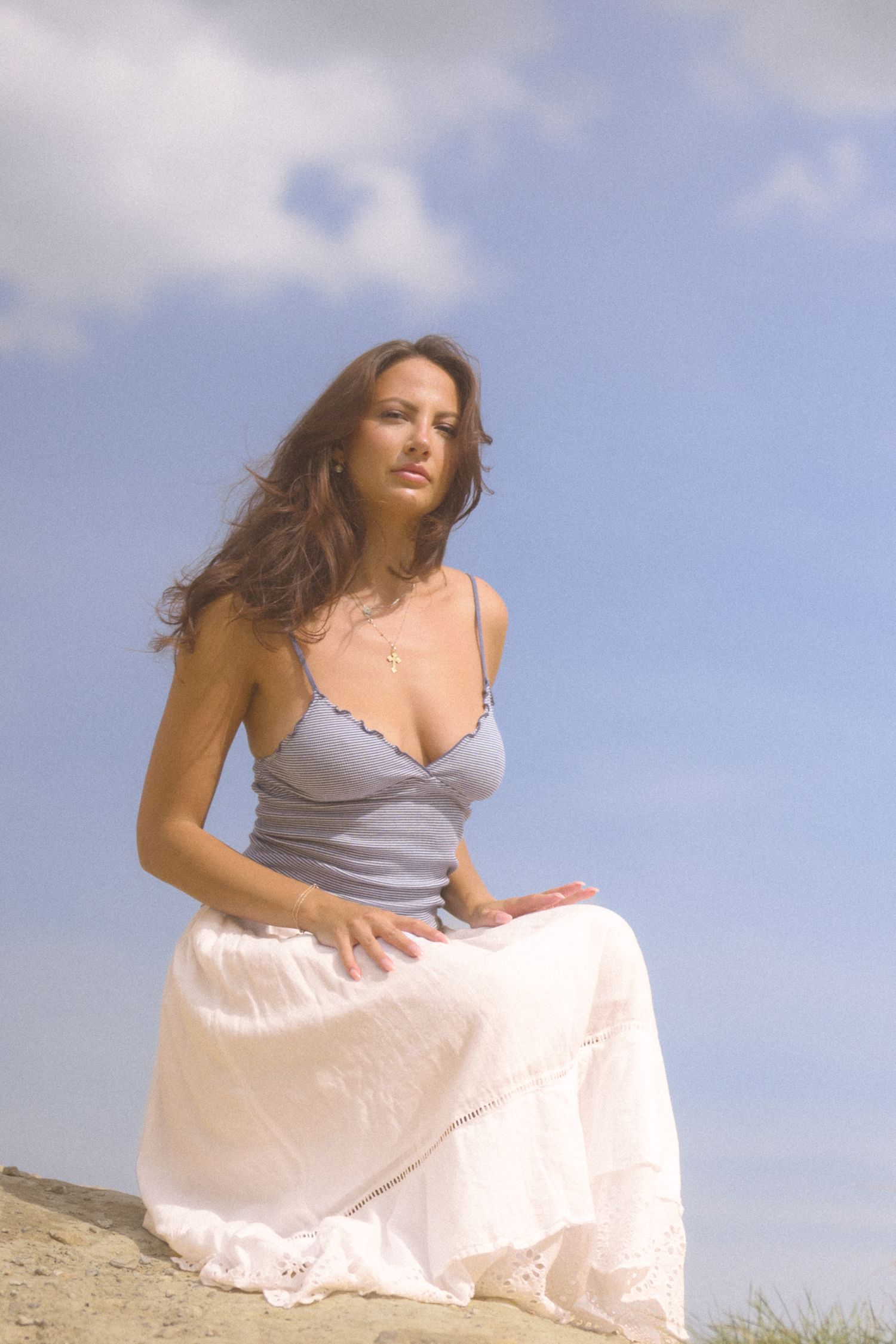 A person in a flowing white skirt and gray top stands outdoors on a sunny day.
