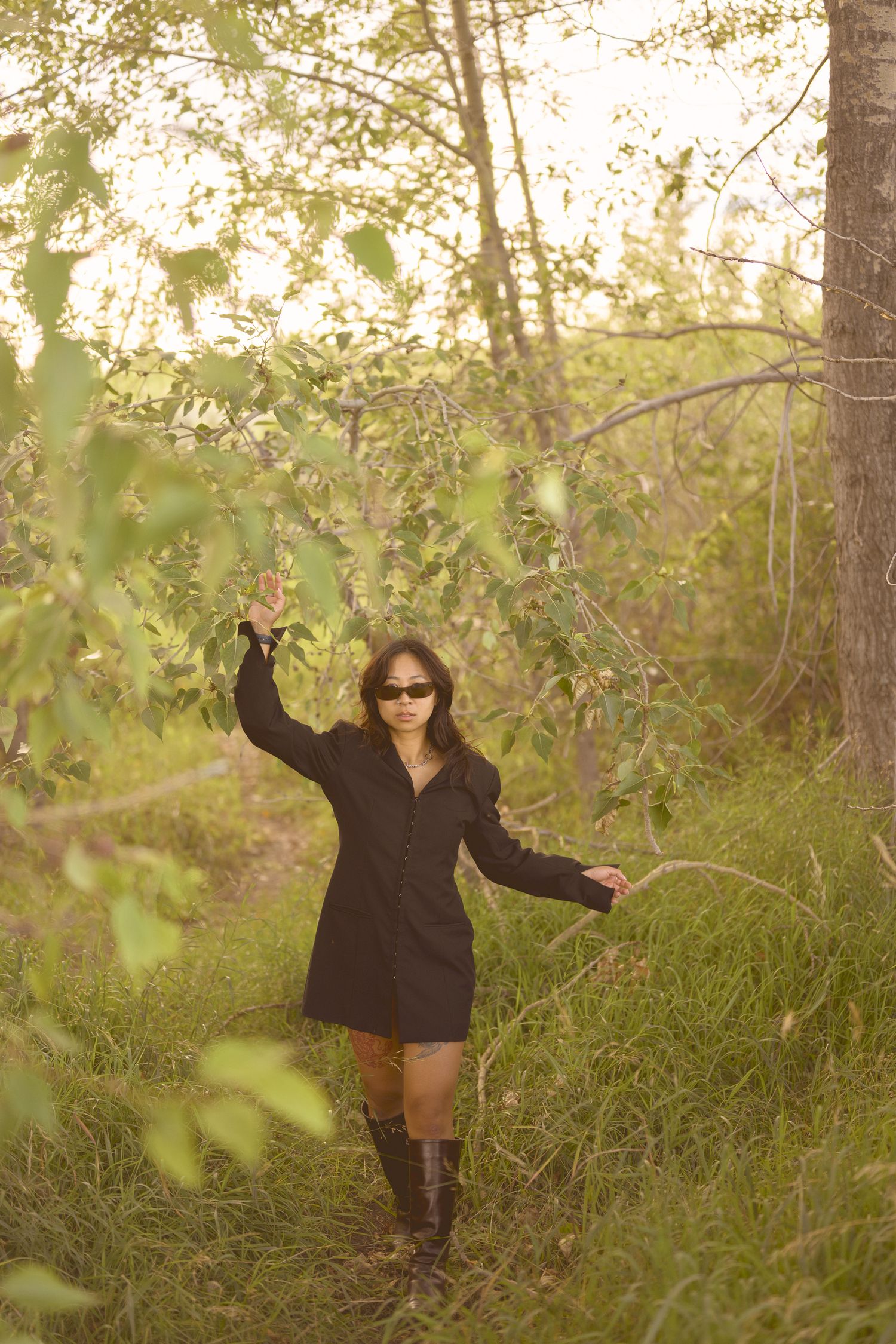 A figure in dark clothing poses playfully among trees and foliage in a wooded area.