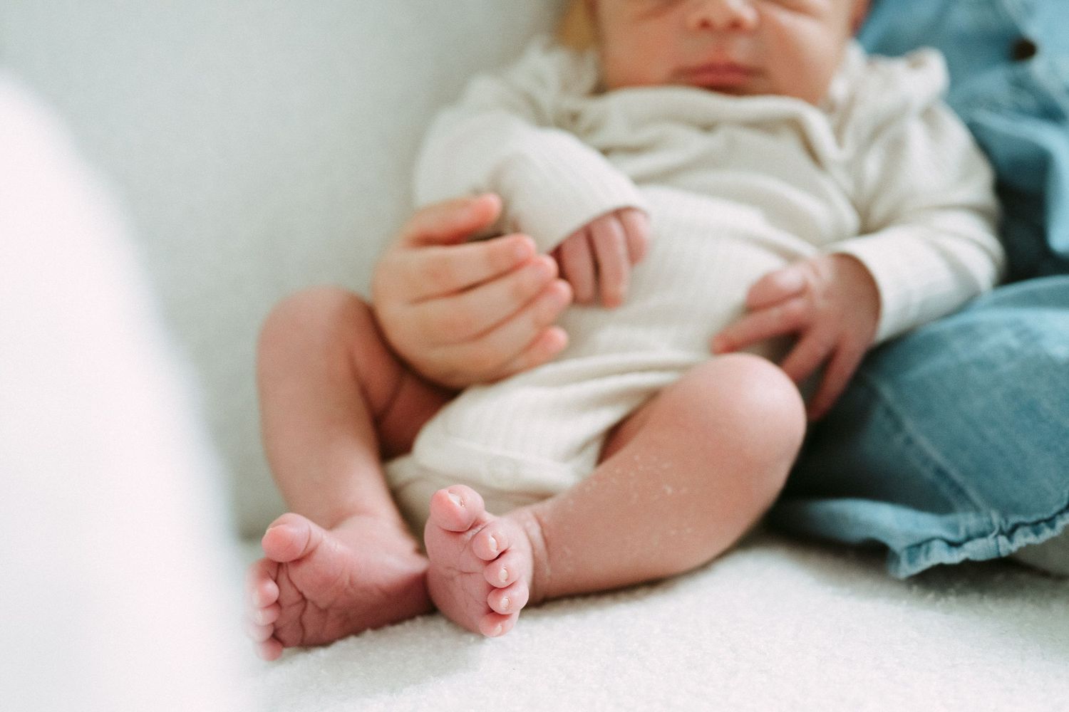 Close-up of newborn baby in white ribbed onesie showing tiny feet and hands resting on soft surface.