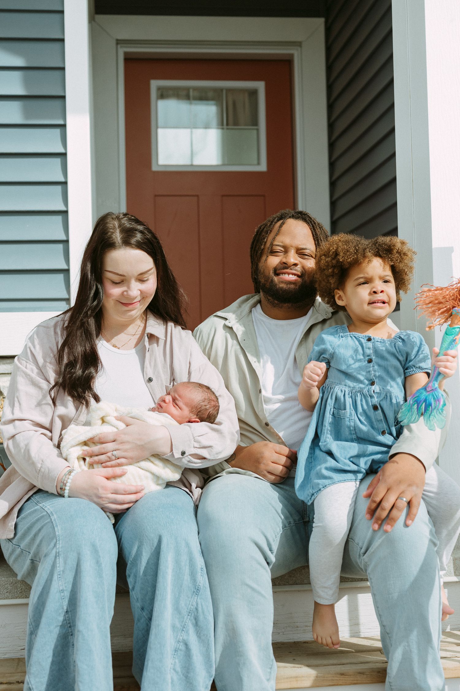 Interracial family of four sitting on front porch steps, mother holding newborn baby beside father and young daughter.