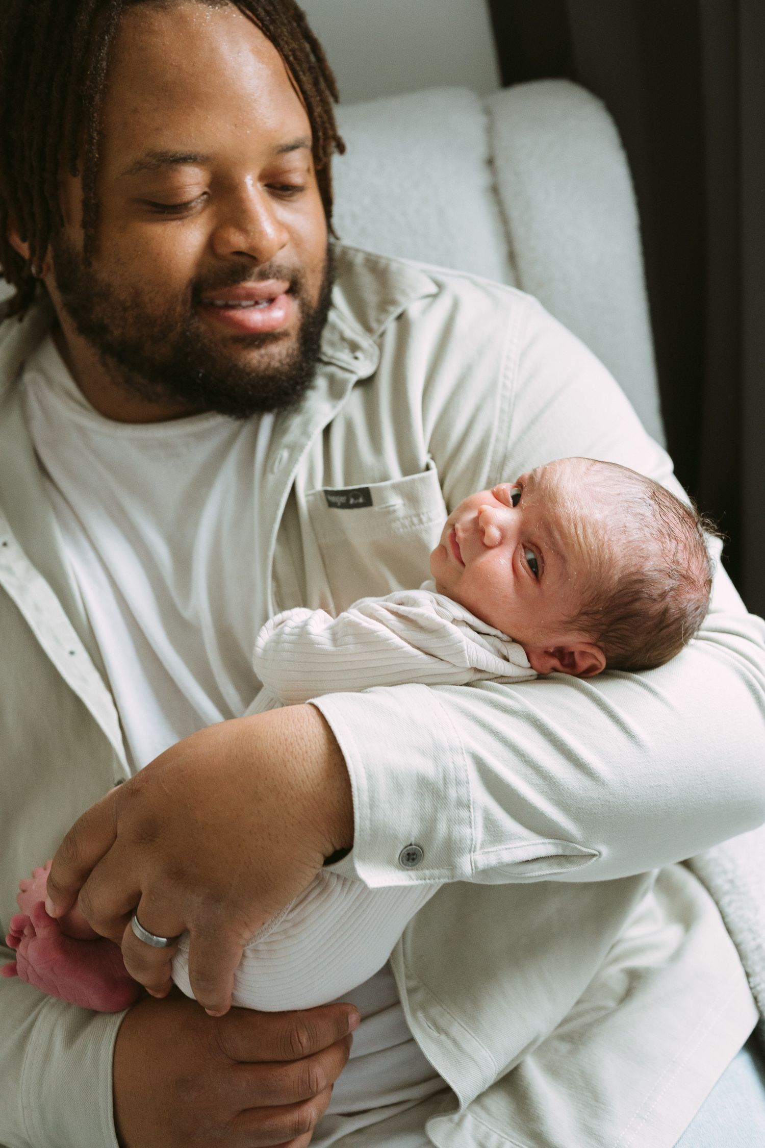 Loving father with dreadlocks cradling newborn baby in his arms while seated in chair.
