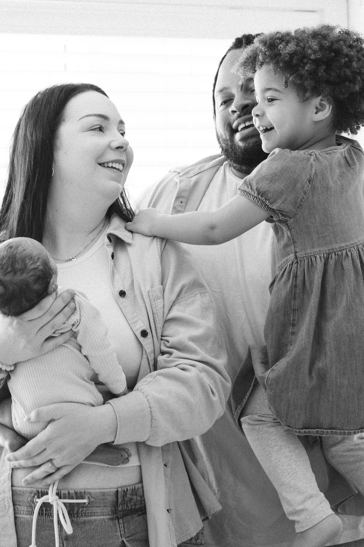 Black and white photo of a happy interracial family with parents holding a newborn and toddler daughter.