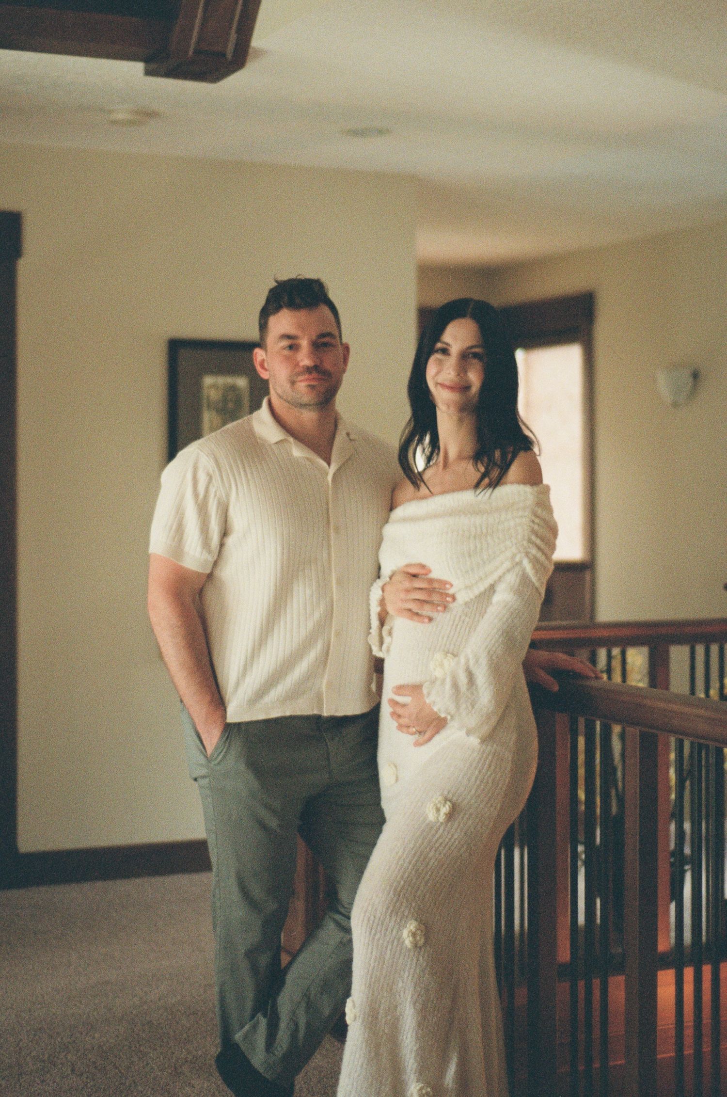 Couple posing indoors for maternity photo shoot, woman in off-shoulder cream knit dress cradling baby bump.