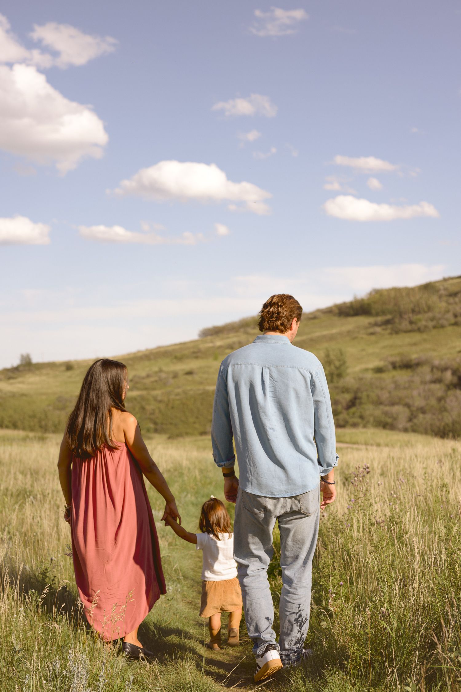 A family walks together through a grassy hillside under a blue sky with fluffy clouds.