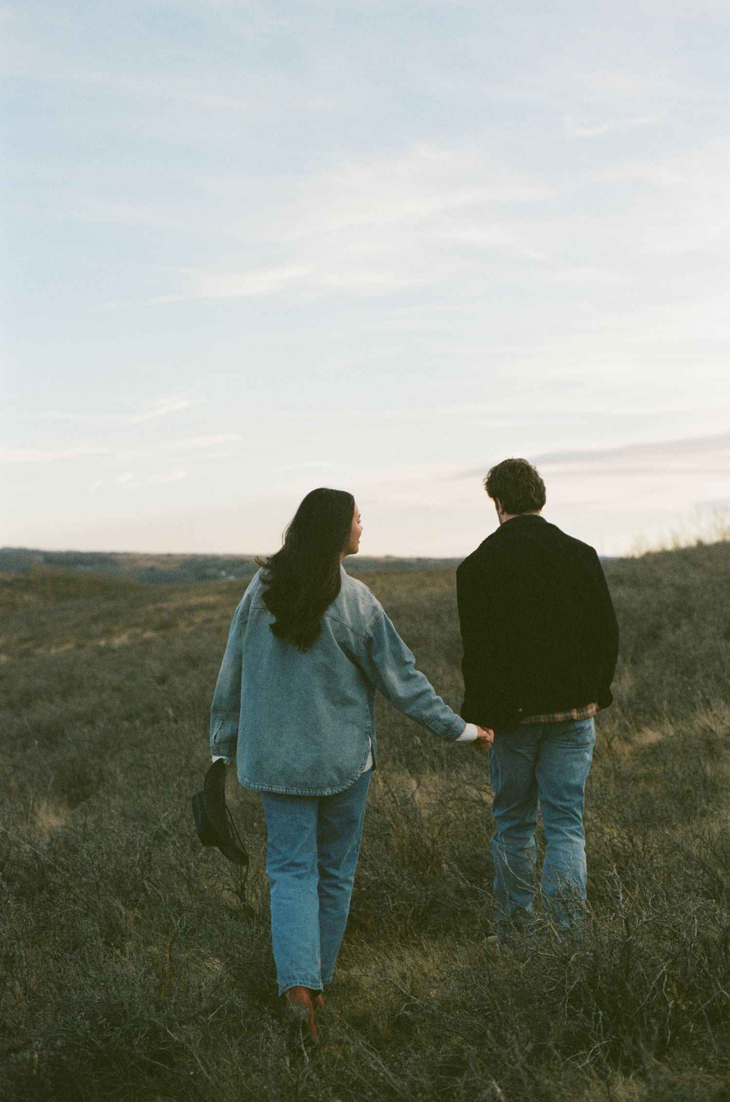 Two people in denim walk together across a grassy hillside during sunset.