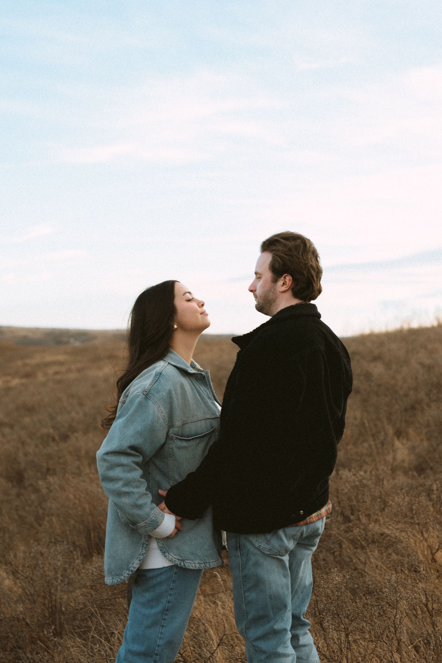 A couple in denim outfits embrace while standing in a brown grassy field against a cloudy sky.