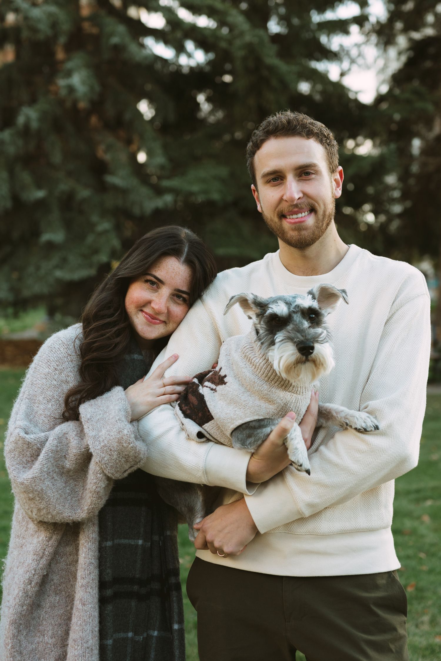 Happy couple holding their small grey dog while posing in a park setting with evergreen trees.