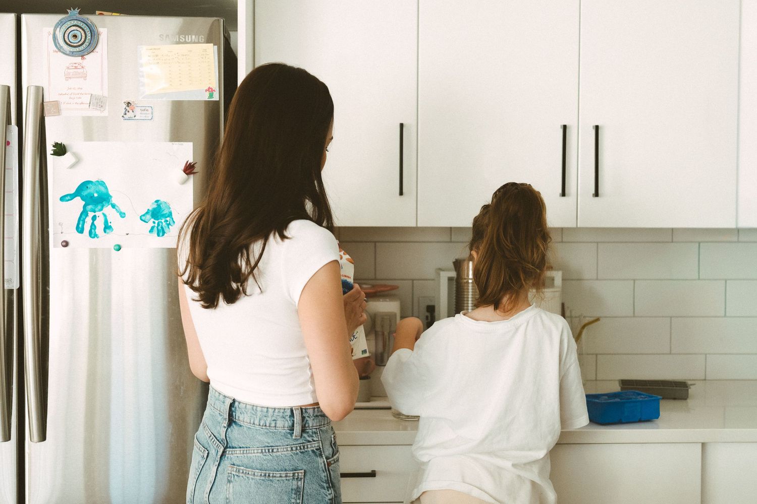 Two people in casual attire stand in a modern white kitchen with subway tile backsplash and white cabinets.