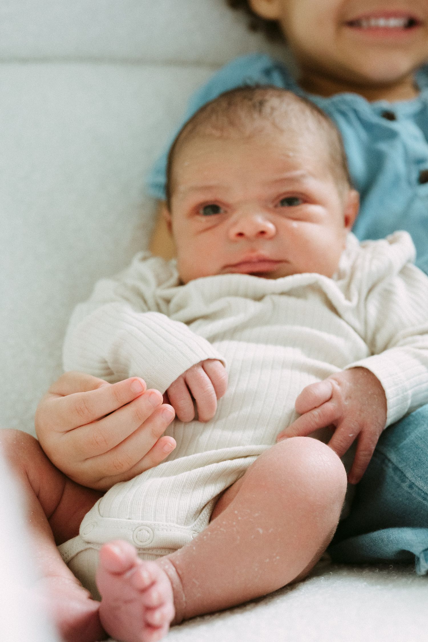 Close-up of newborn baby in cream onesie being held by smiling older sibling on white chair.