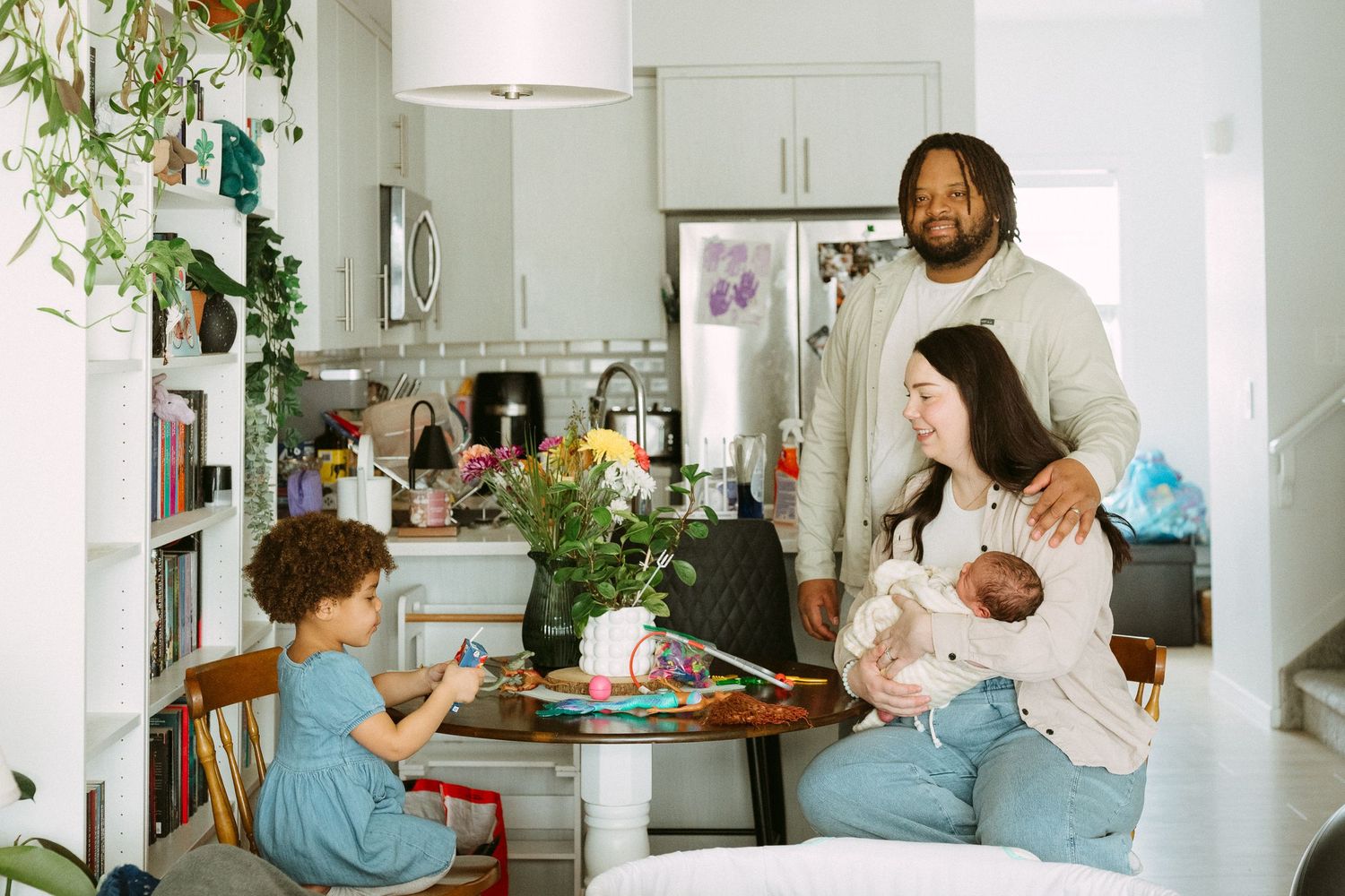 Family of four at home, parents with newborn baby and toddler gathered around dining table in bright kitchen.