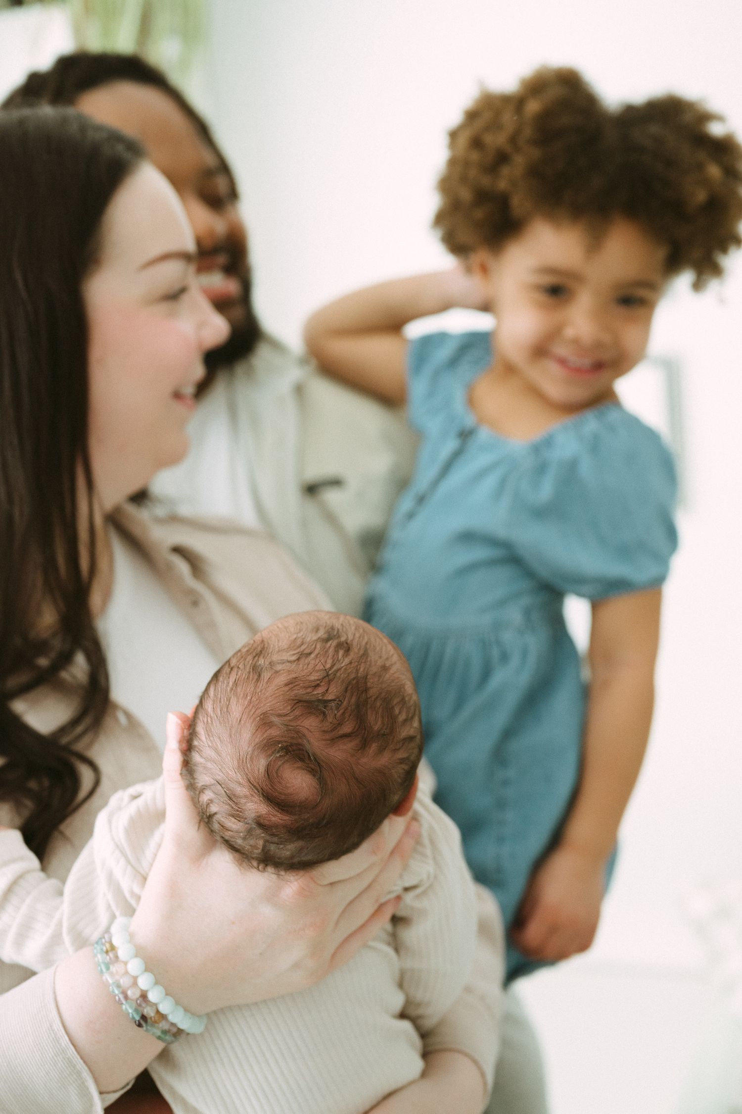 Close-up of a mother cradling a newborn baby with curly-haired toddler smiling in the background.