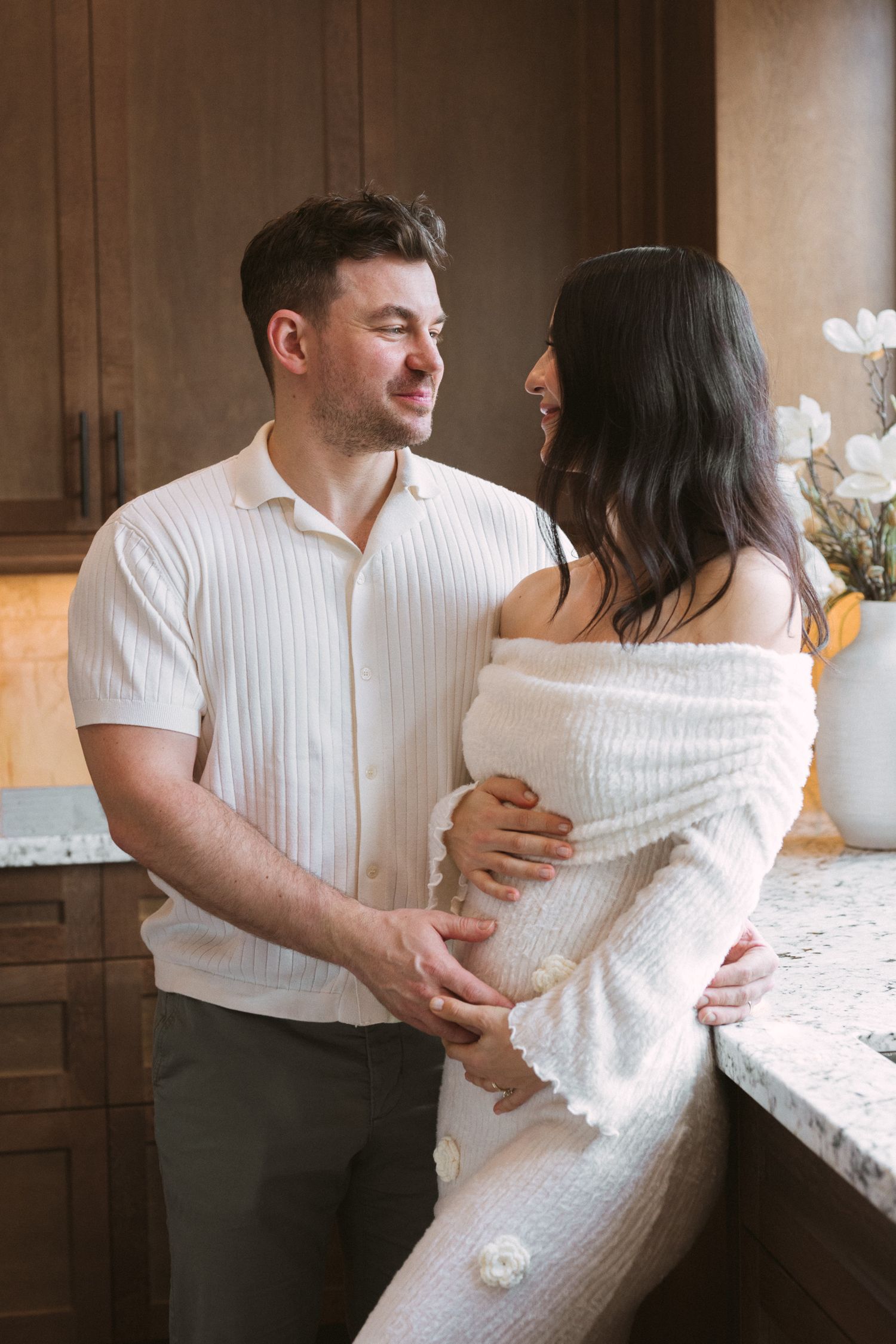A couple in white outfits share a tender moment in a kitchen, with the woman showing a baby bump.