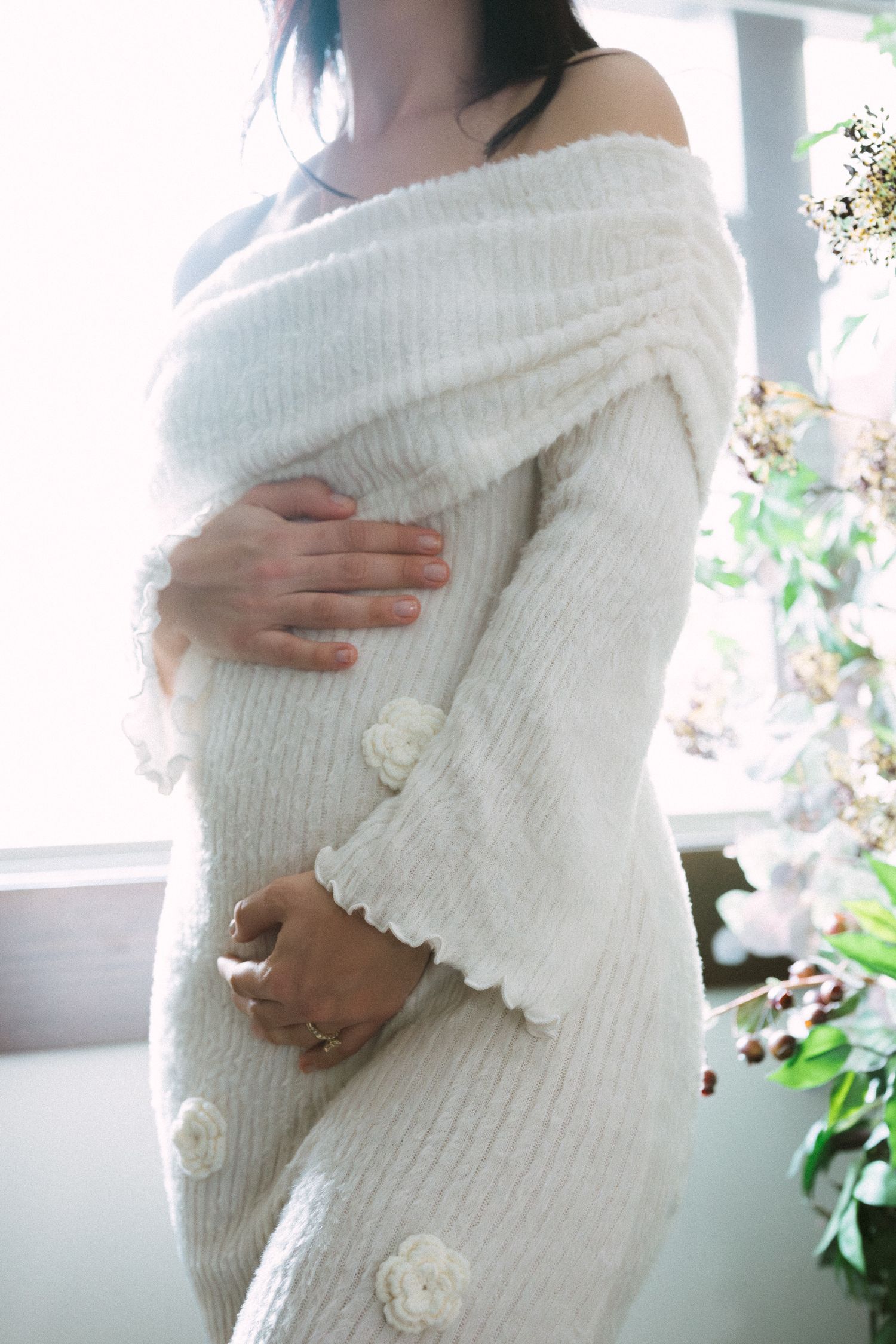Close-up of a pregnant woman in a white off-shoulder knit dress cradling her baby bump.