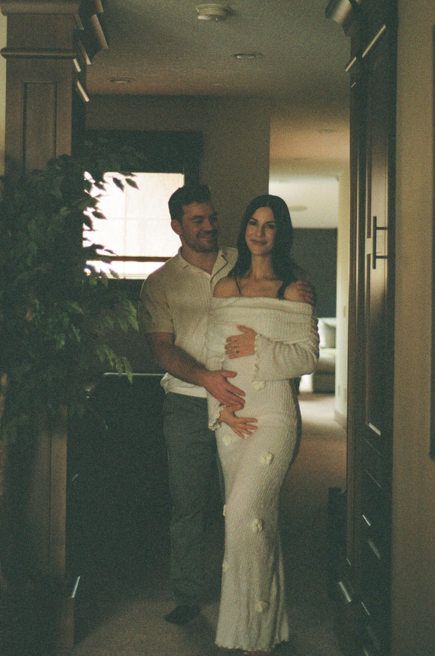 Couple embraces in hallway, pregnant woman in white floral dress with partner in cream shirt.