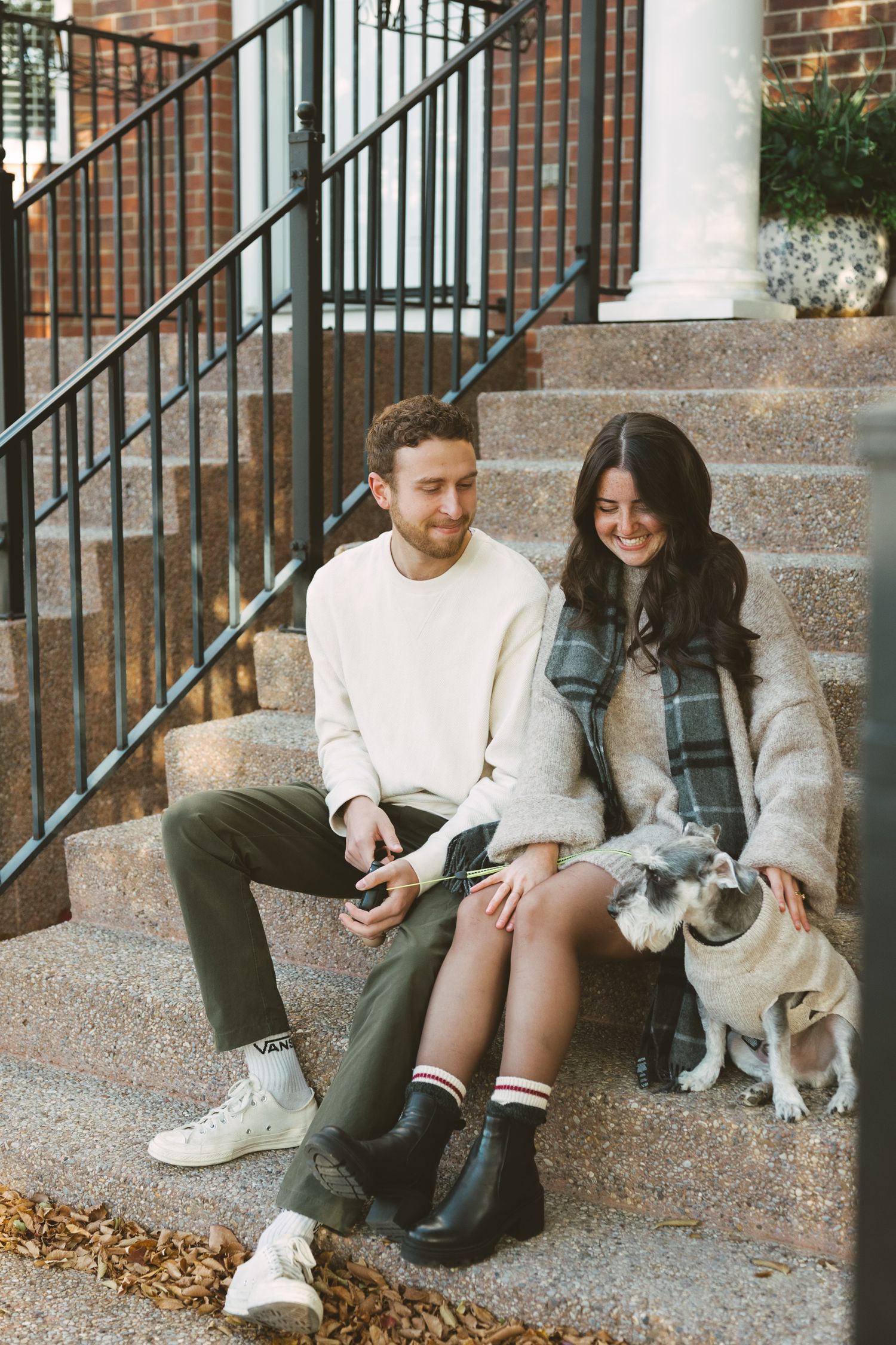 A couple sits together on brick steps with their small dog, wearing casual fall attire in an urban setting.