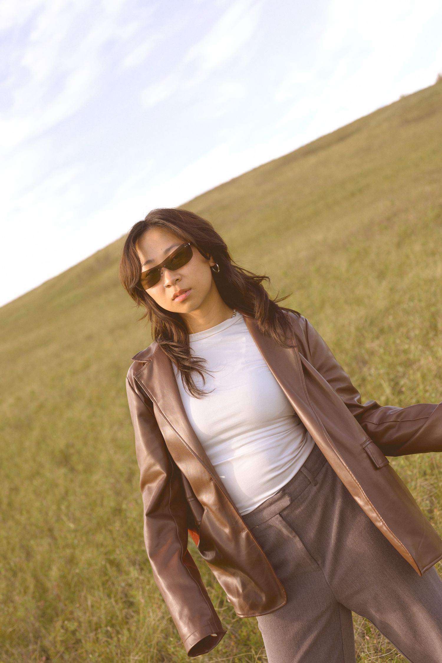 A figure in a brown leather jacket and white top poses against a grassy hillside.
