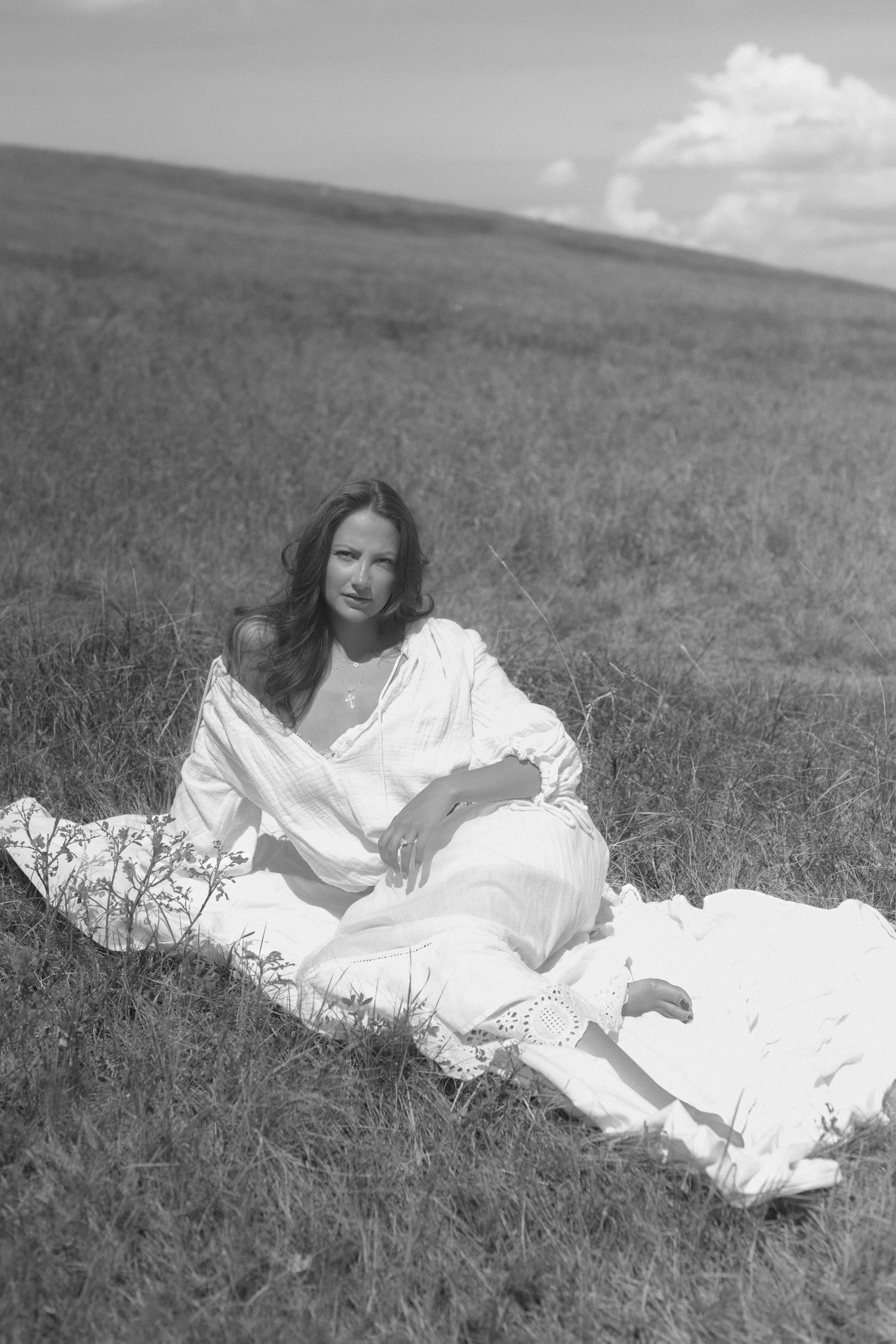 Black and white portrait of someone reclining in a white dress on a grassy hillside.