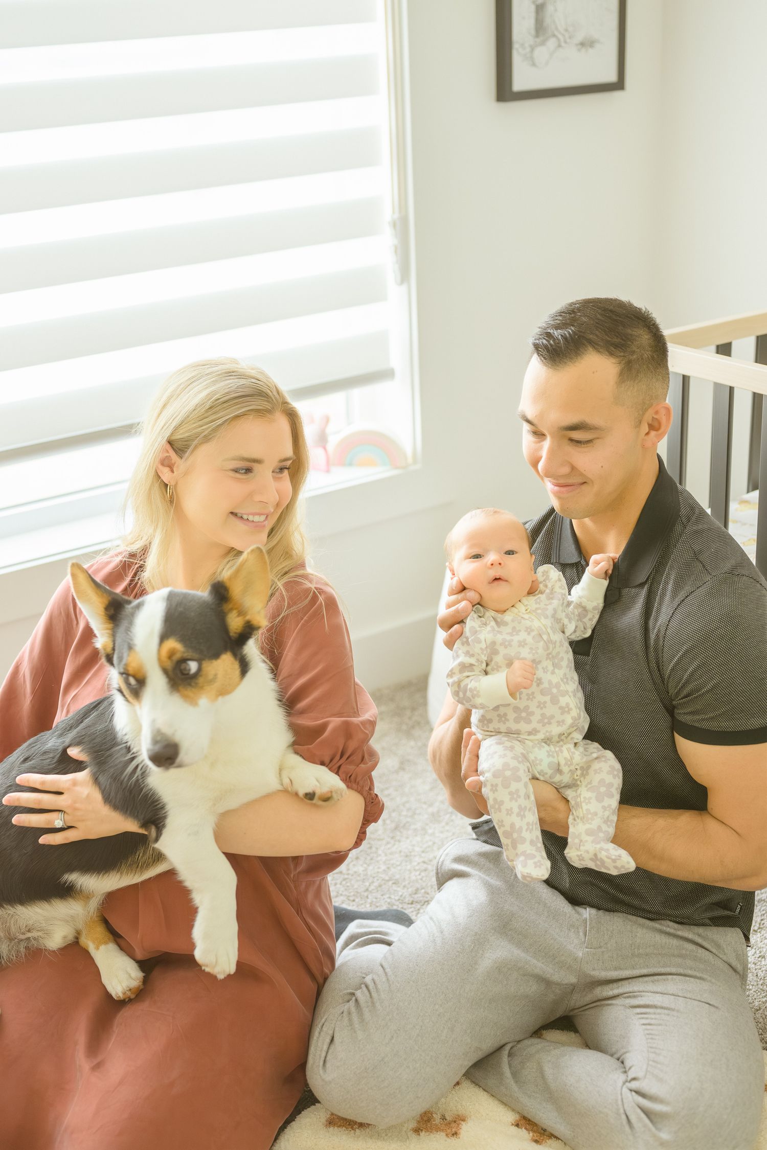 A couple sits together with their infant and tricolor dog in a bright nursery setting.