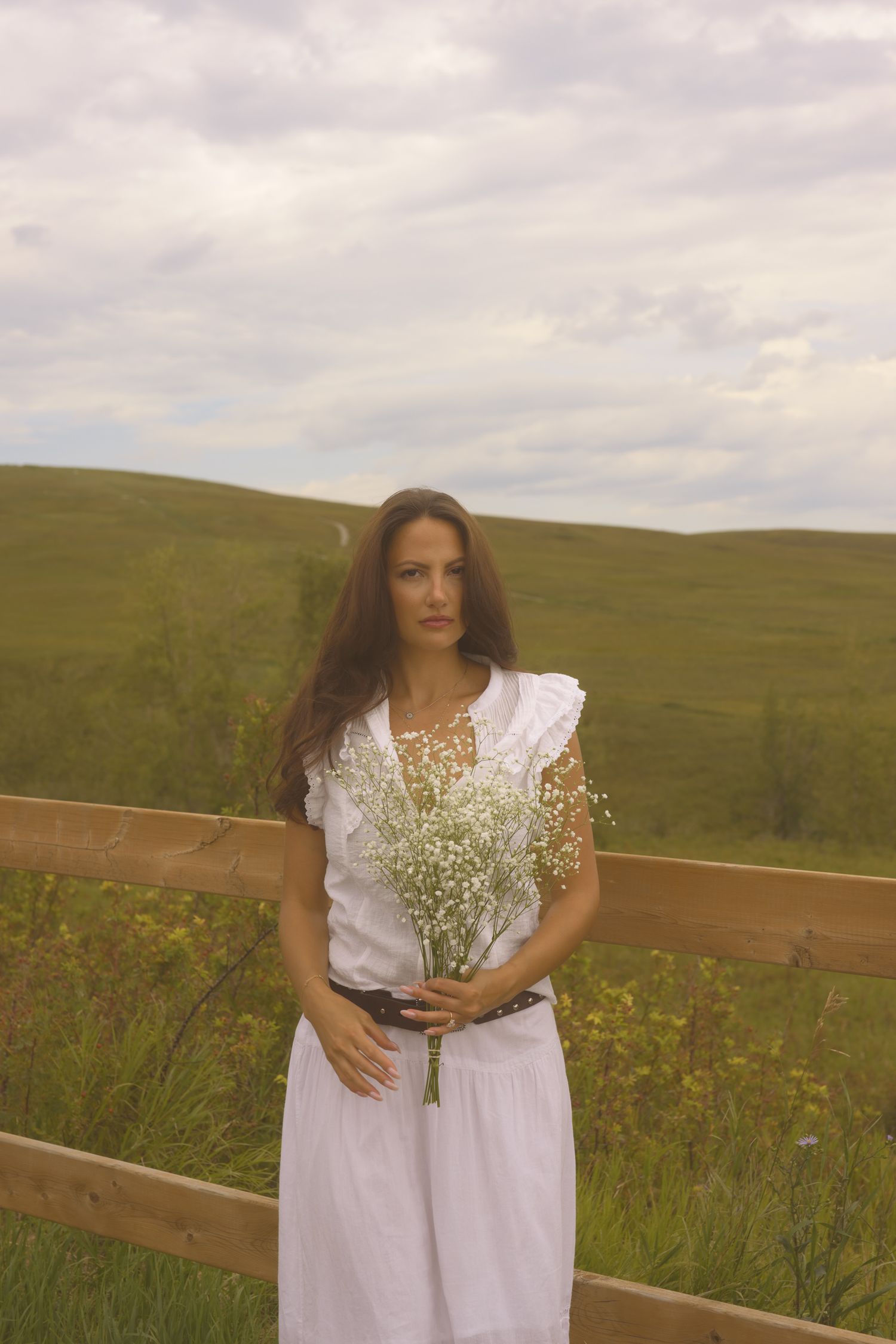 Person in white dress holds bouquet of baby's breath flowers against rural hillside backdrop.