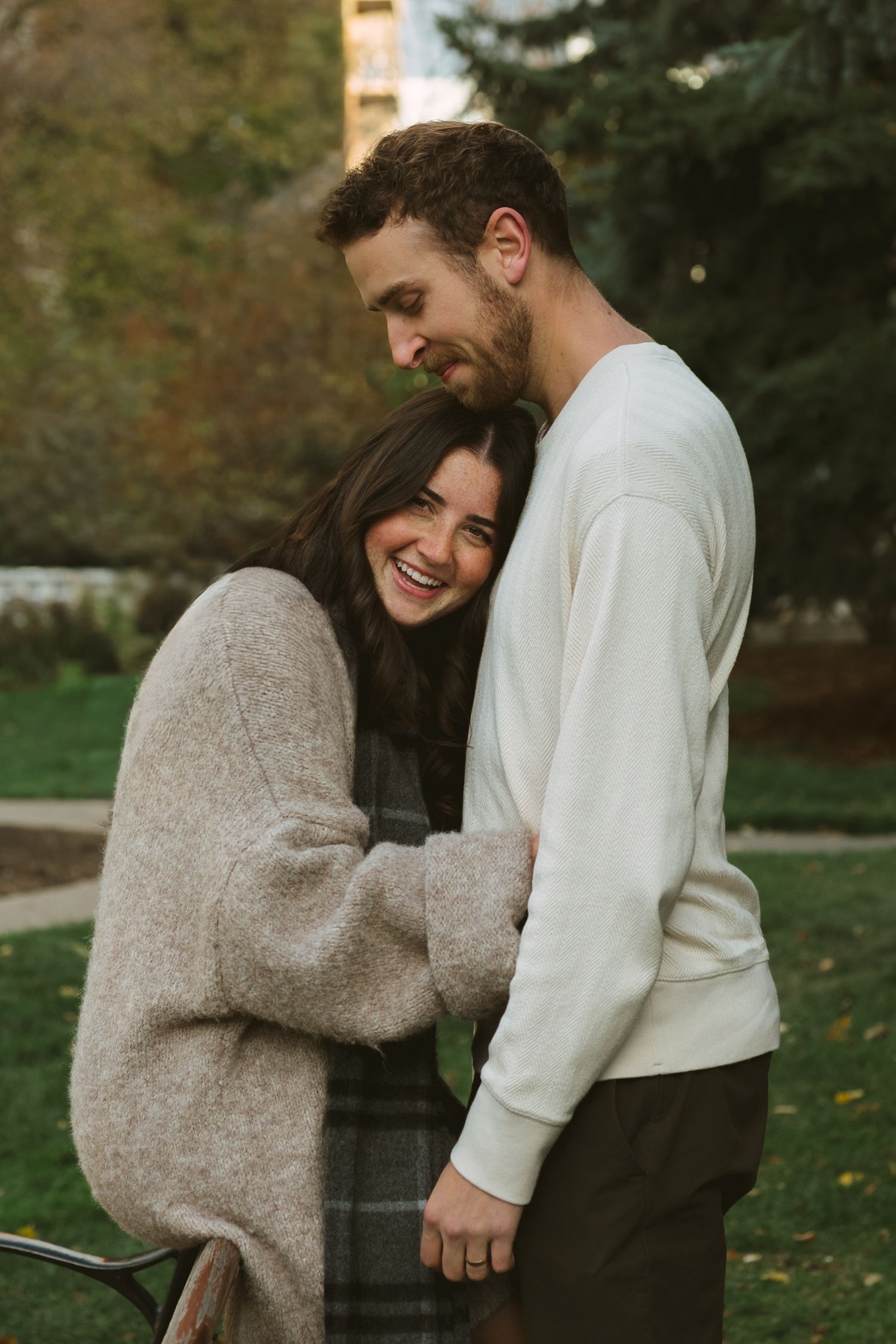 A couple in autumn attire share intimate moments in a park setting with fallen leaves.