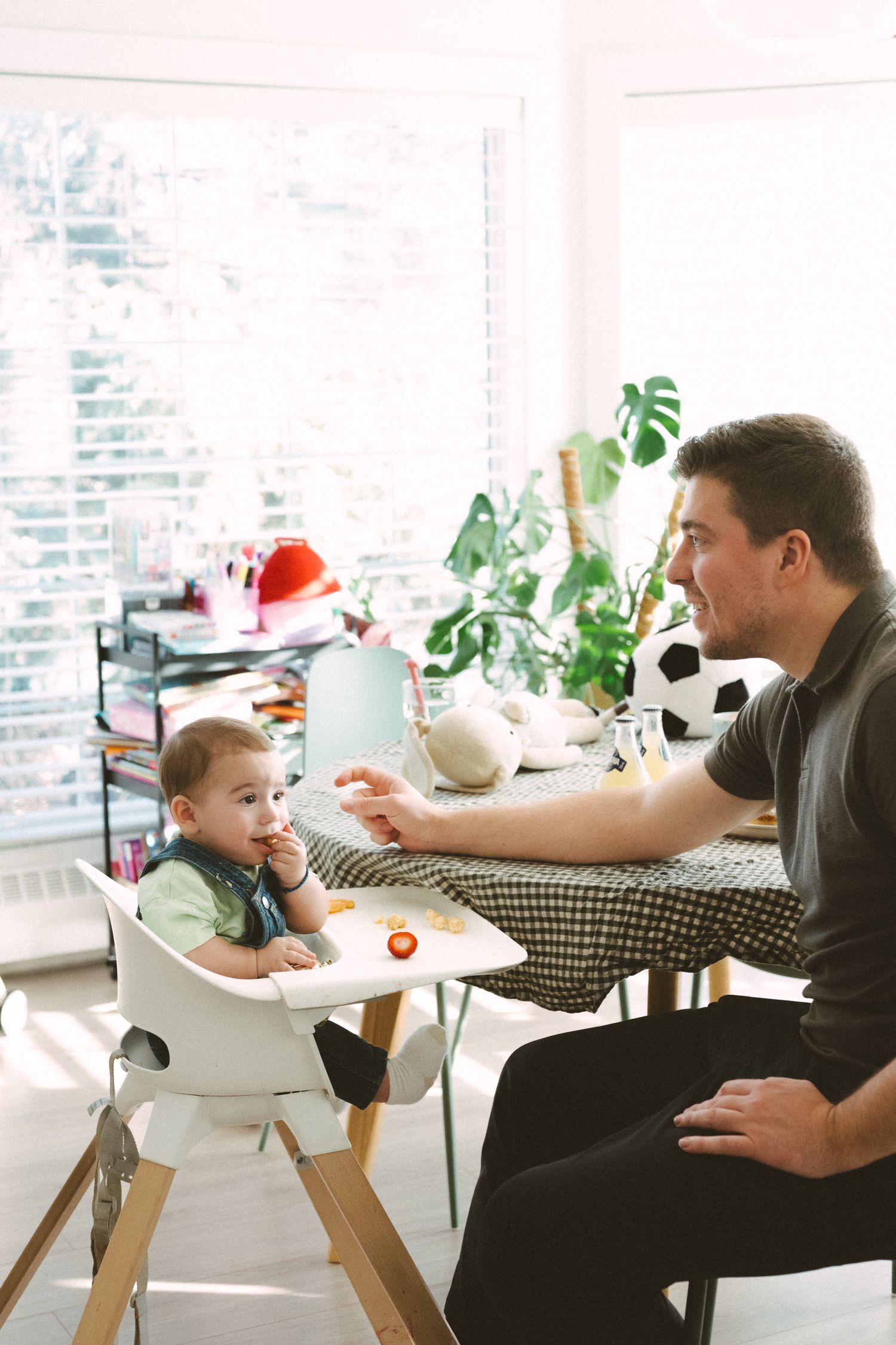 Family mealtime scene in bright kitchen with high chair, houseplants and natural light streaming through windows.