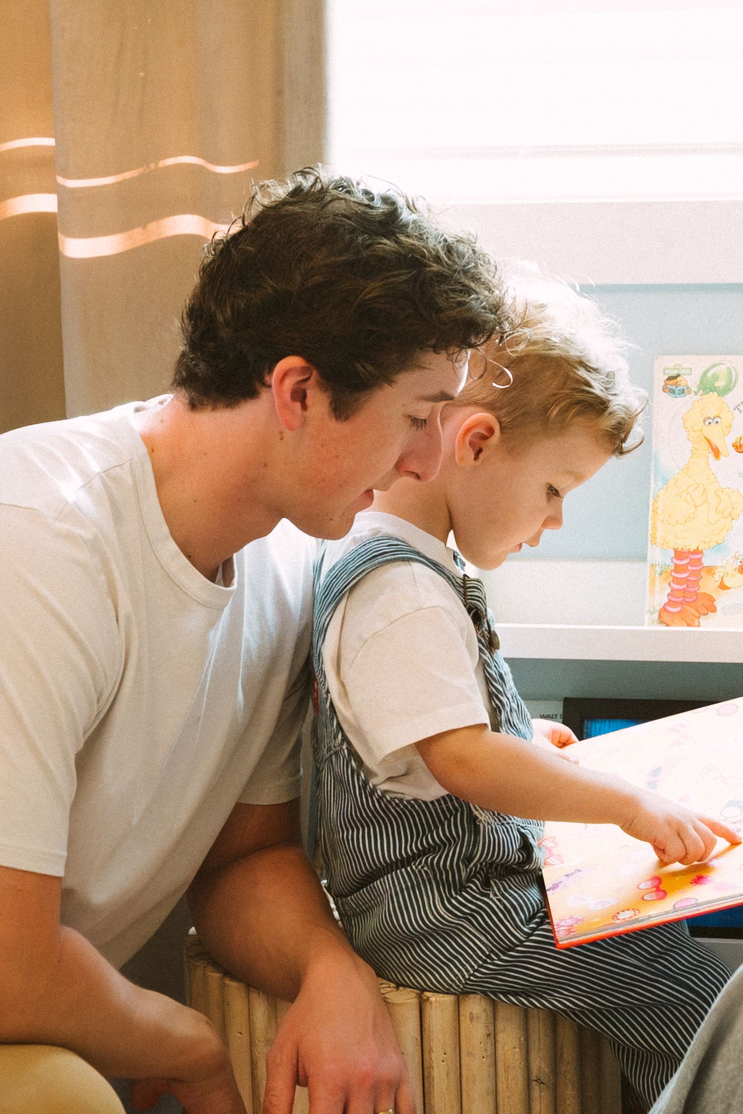 A child in striped overalls reads a book while sitting with an adult in a white t-shirt.