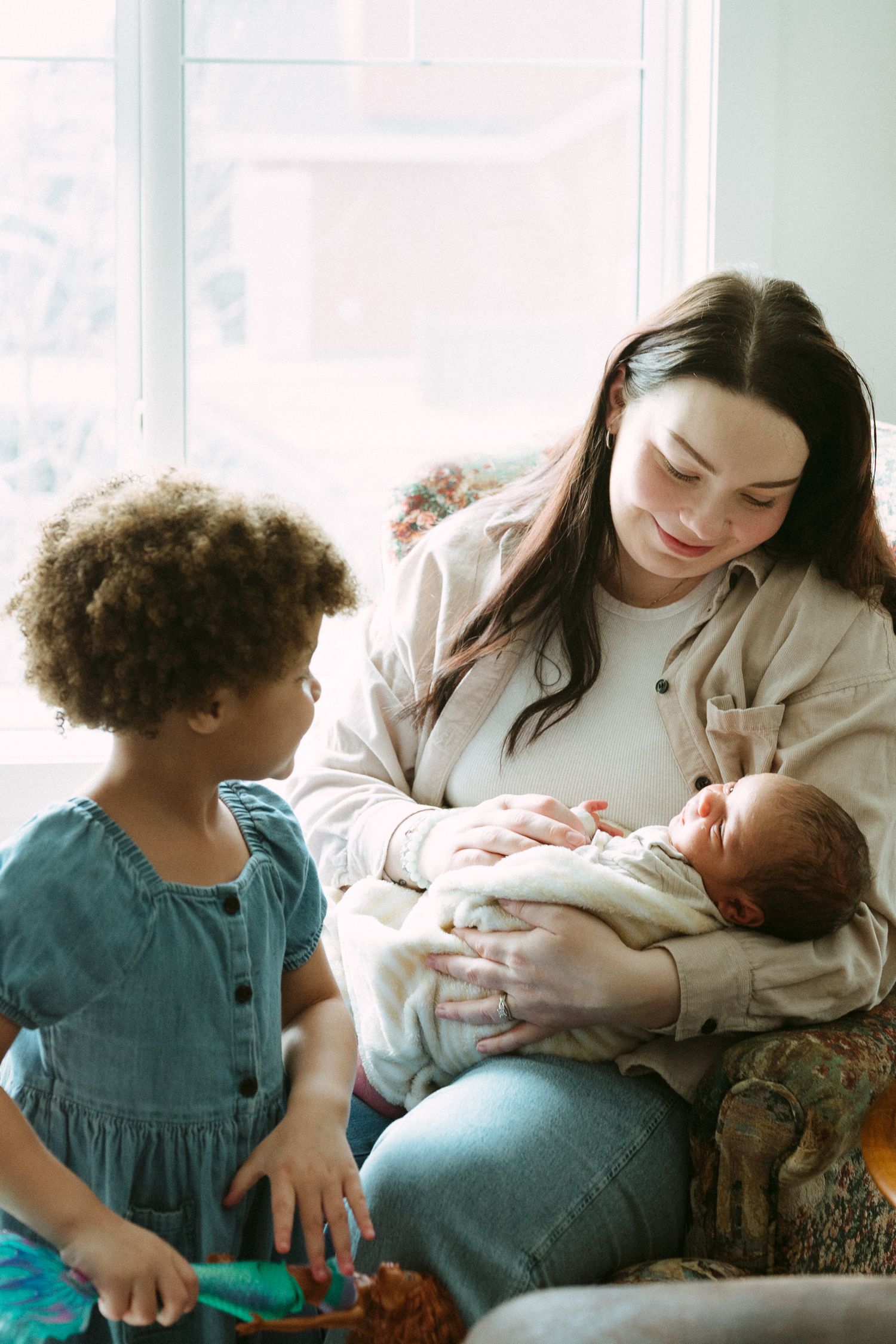 A mother smiles while cradling her newborn baby as her young curly-haired toddler looks on with curiosity.