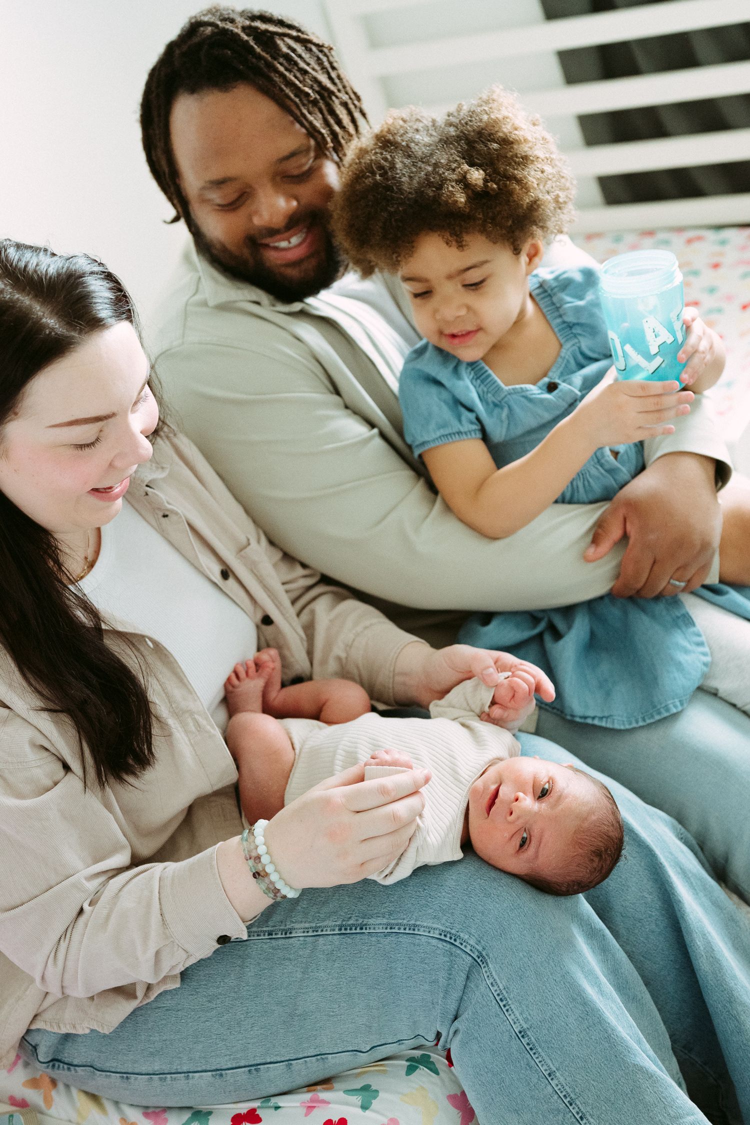 Multiracial family sitting together on a bed, parents smiling at newborn baby while toddler holds a book.