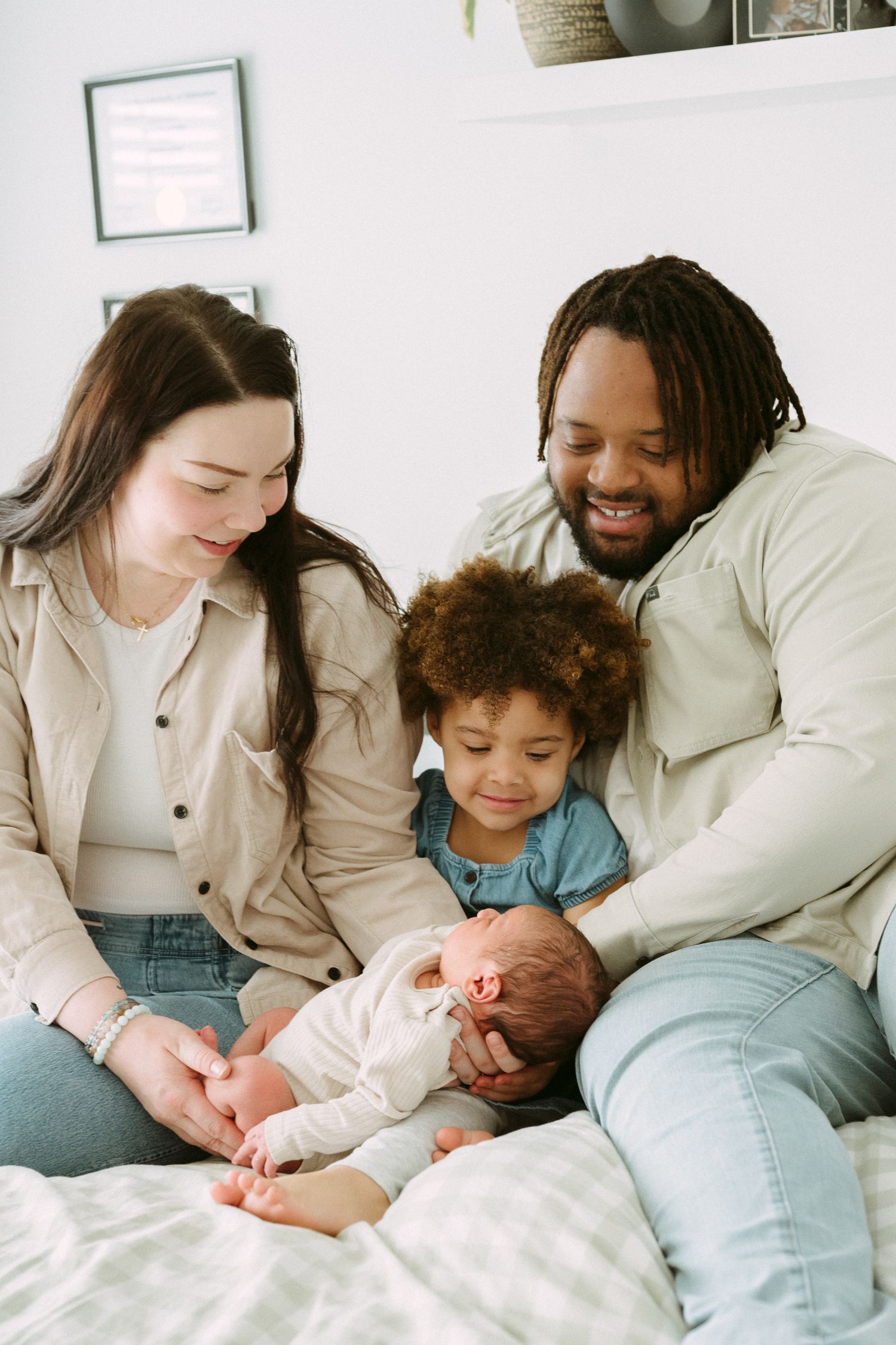 Happy interracial family of four sitting on bed, parents smiling at newborn baby while toddler looks on.