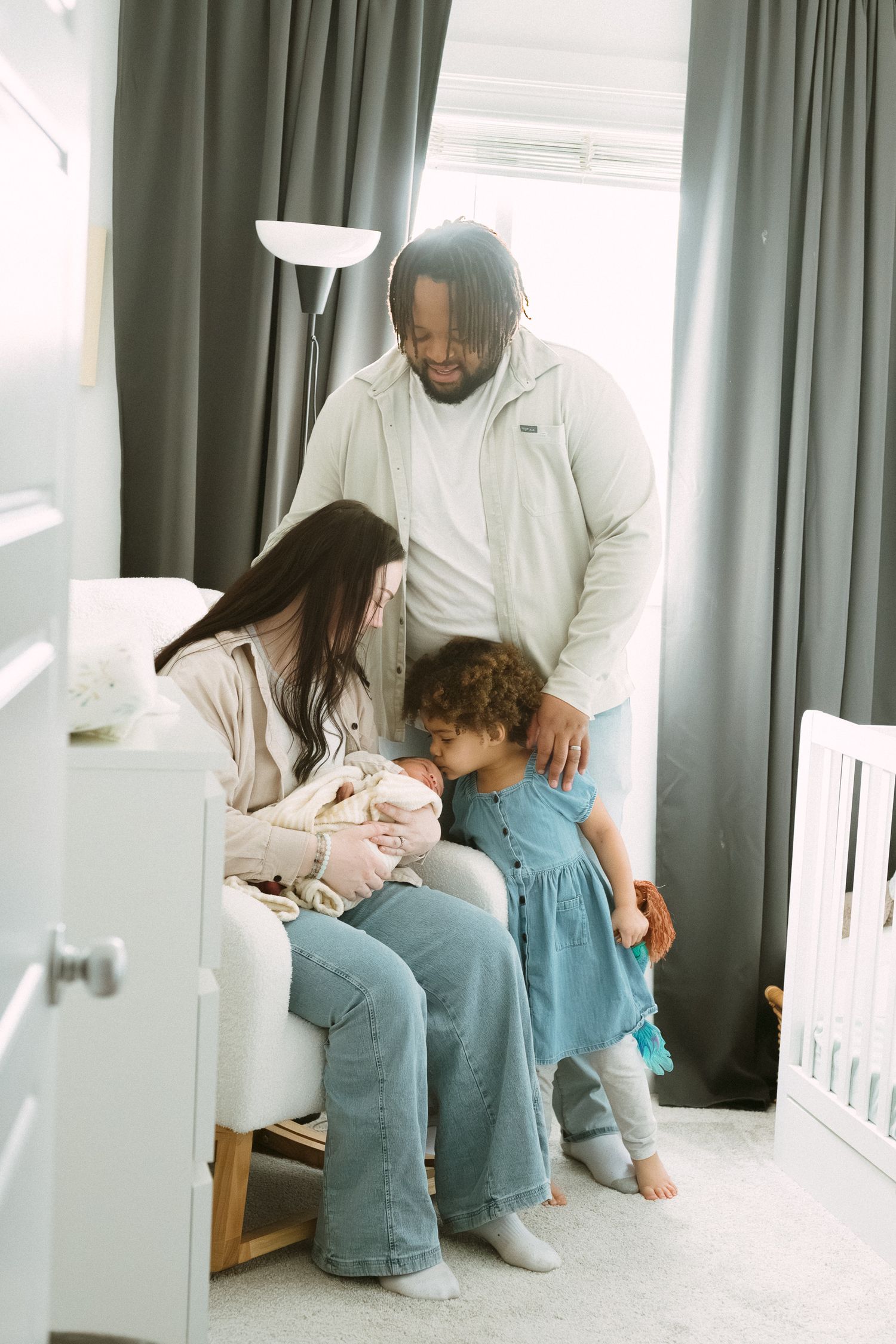 A loving multiracial family gathers around their newborn in a bright nursery, with toddler leaning in for a closer look.