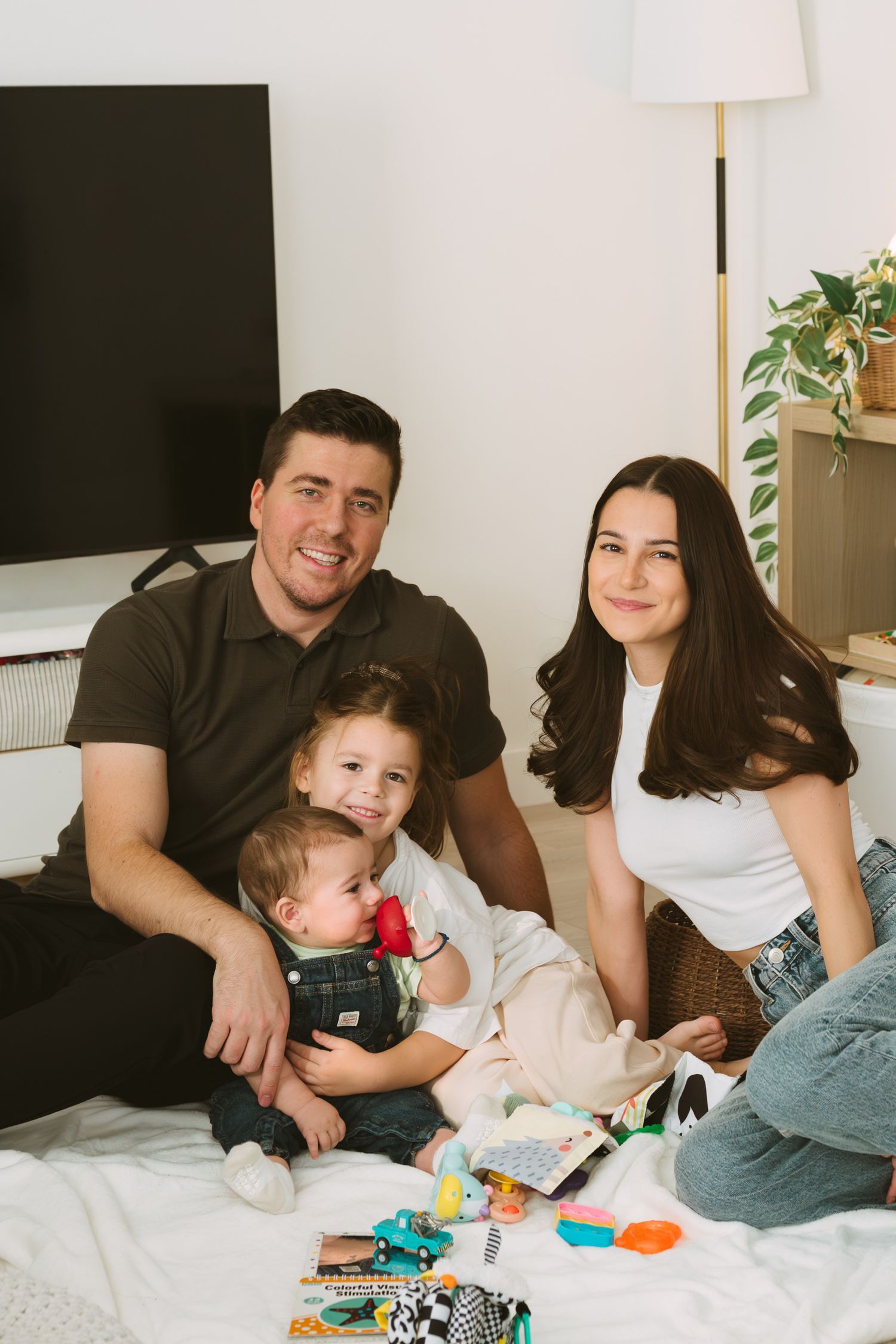 Family lounging on white bed playing with colorful toys together in bright modern bedroom.