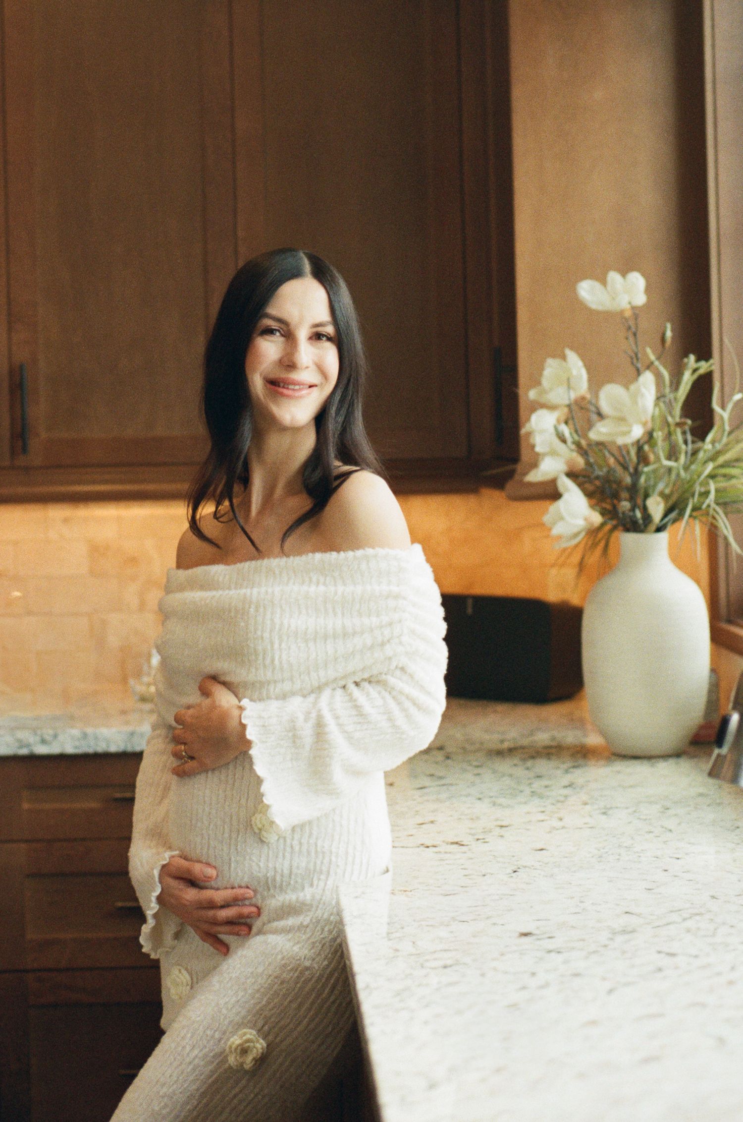 Happy pregnant woman in white off-shoulder dress leaning on kitchen counter beside elegant white vase.