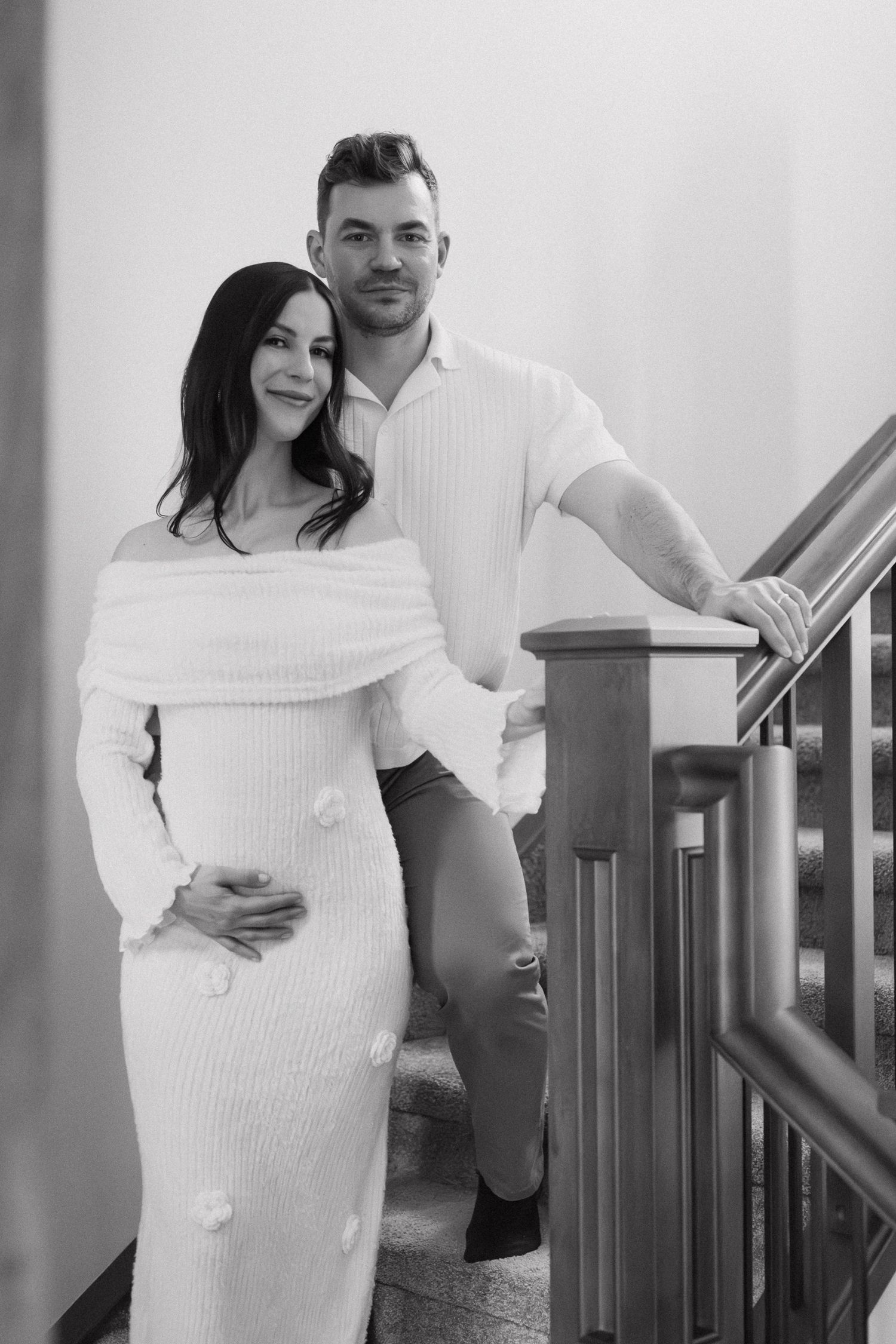 Black and white photo of pregnant couple on staircase, woman in white knit maternity dress holding baby bump.