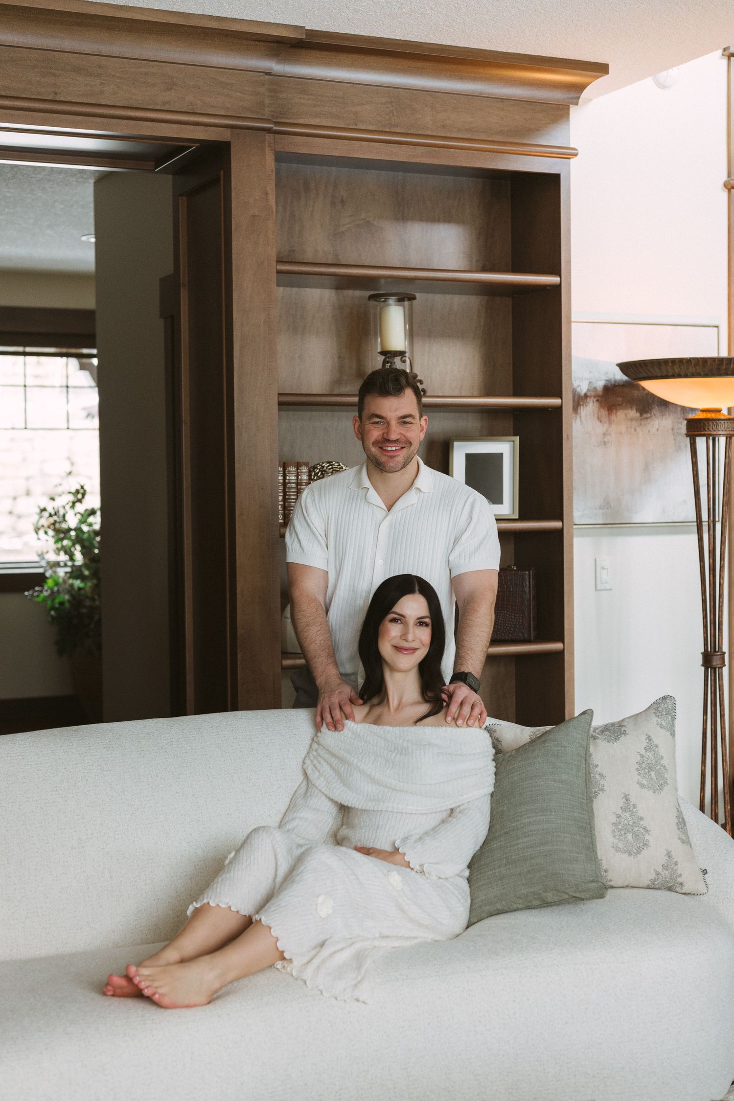 Smiling couple in white outfits posing in a cozy living room with wooden shelving.