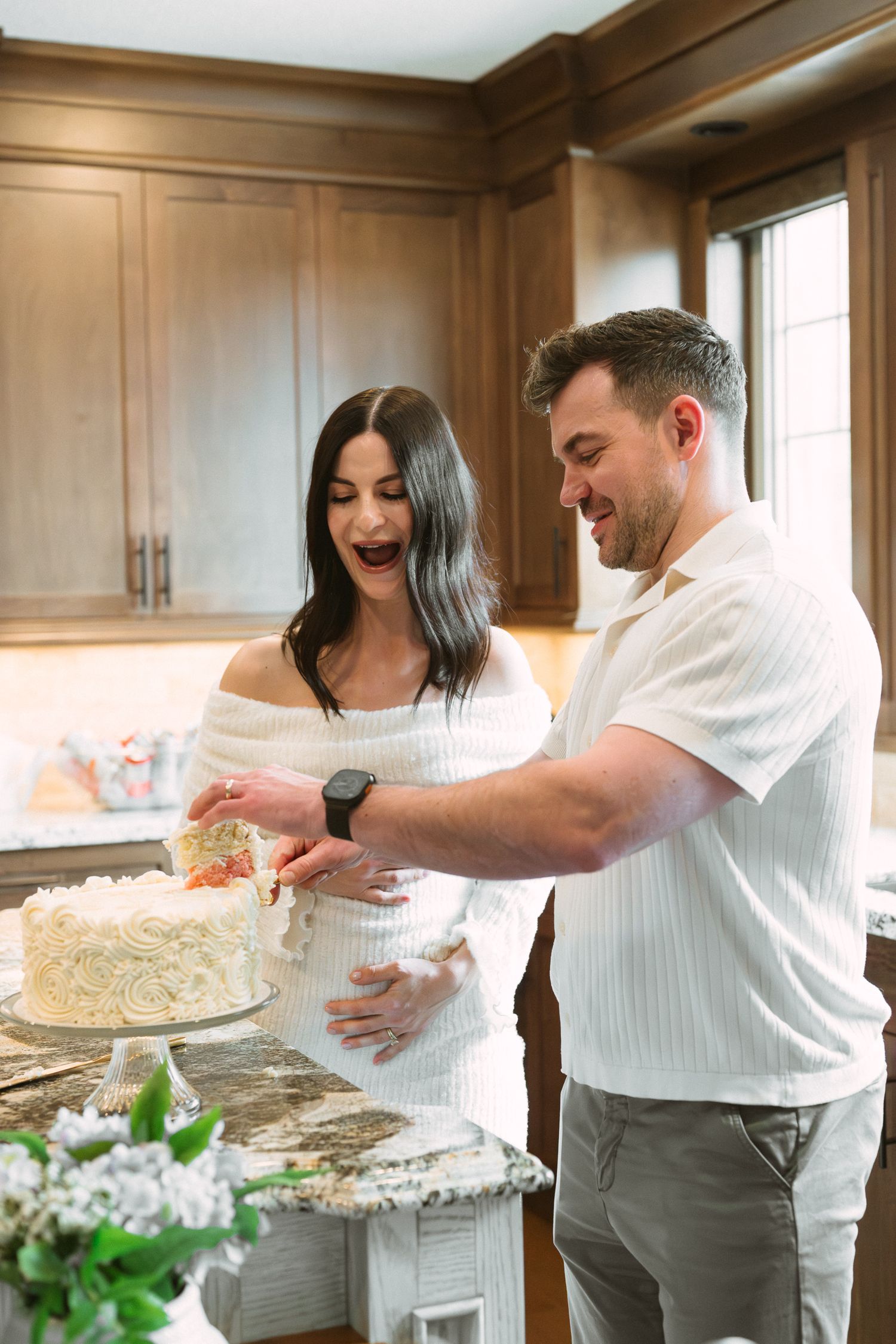 Smiling couple cuts baby shower cake together in kitchen, pregnant woman in white dress with partner.