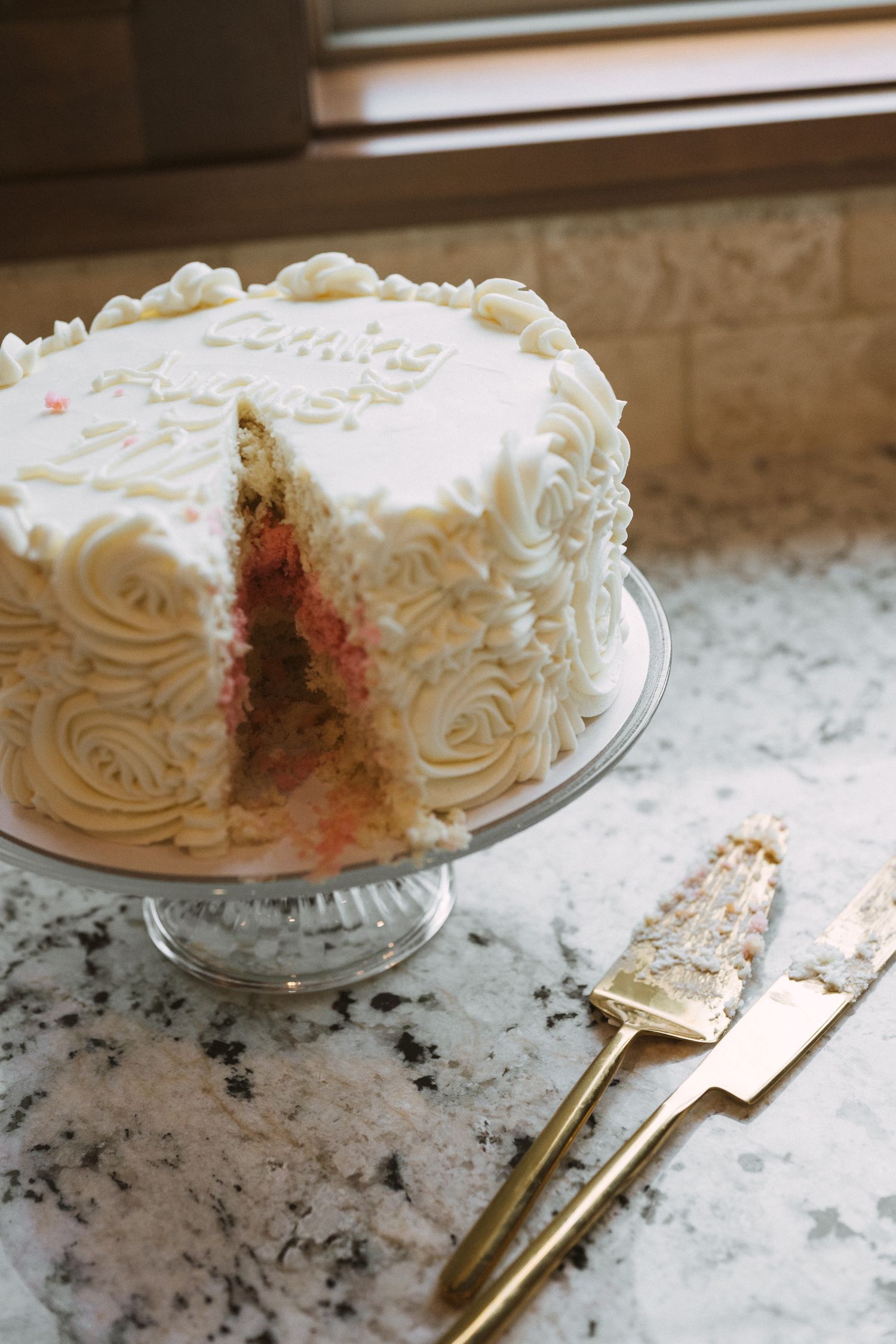 White frosted celebration cake with rosette piping on glass stand, slice removed, gold serving utensils on marble counter.