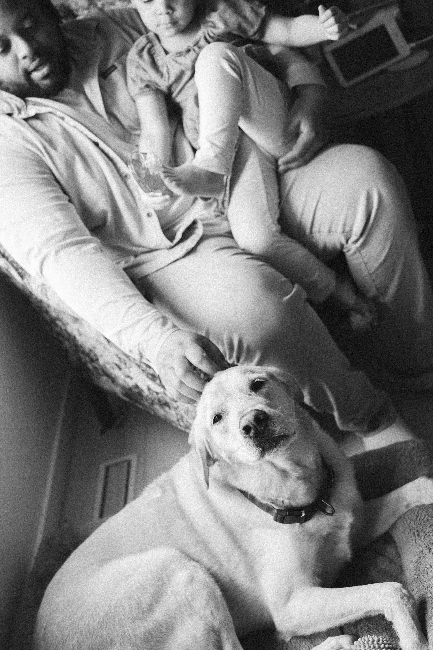 Black and white photo of a person sitting on a couch holding a baby, with a Labrador dog resting nearby.