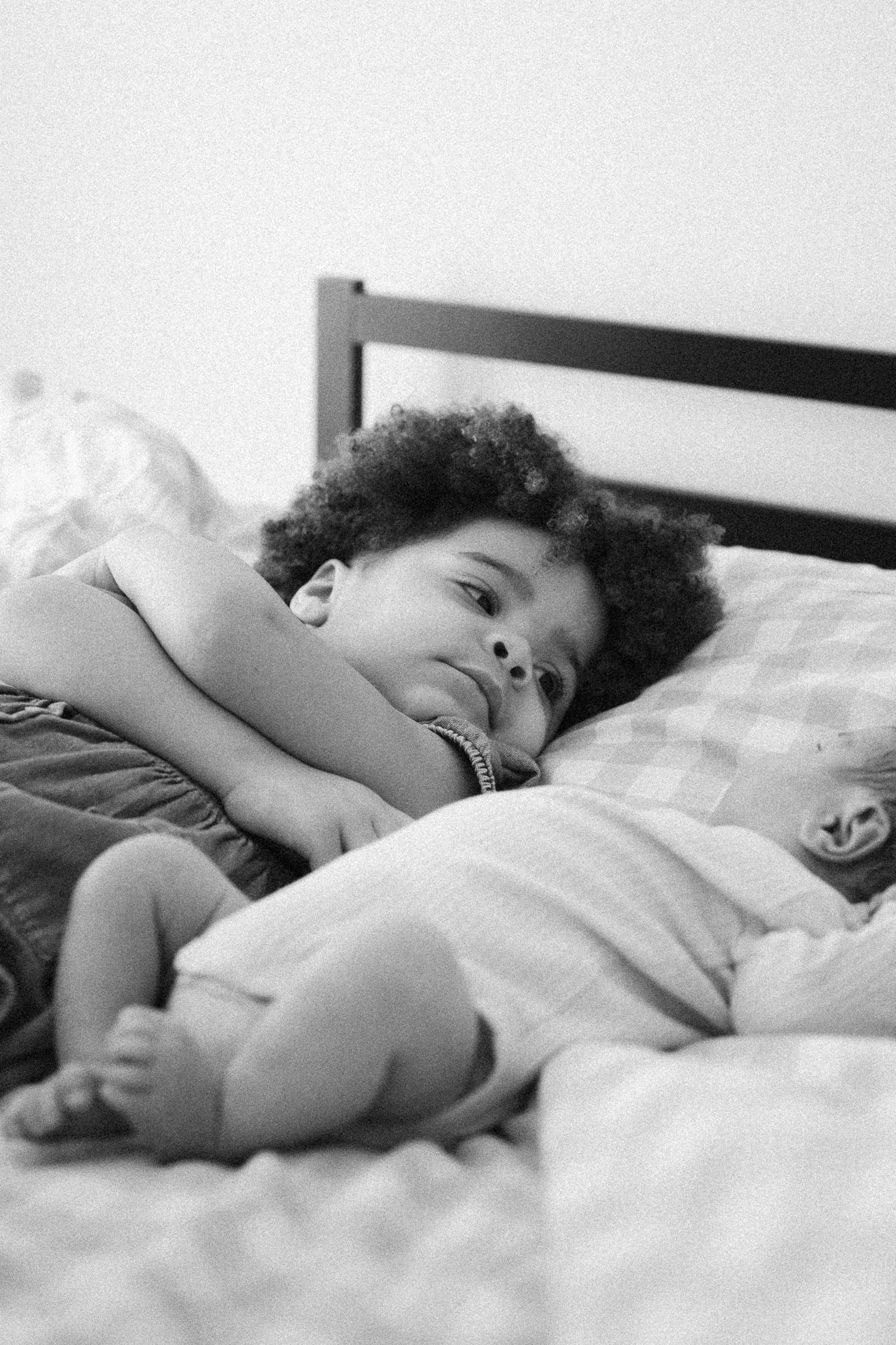 Black and white photo of a curly-haired toddler lying on a bed smiling, with a newborn baby beside them.