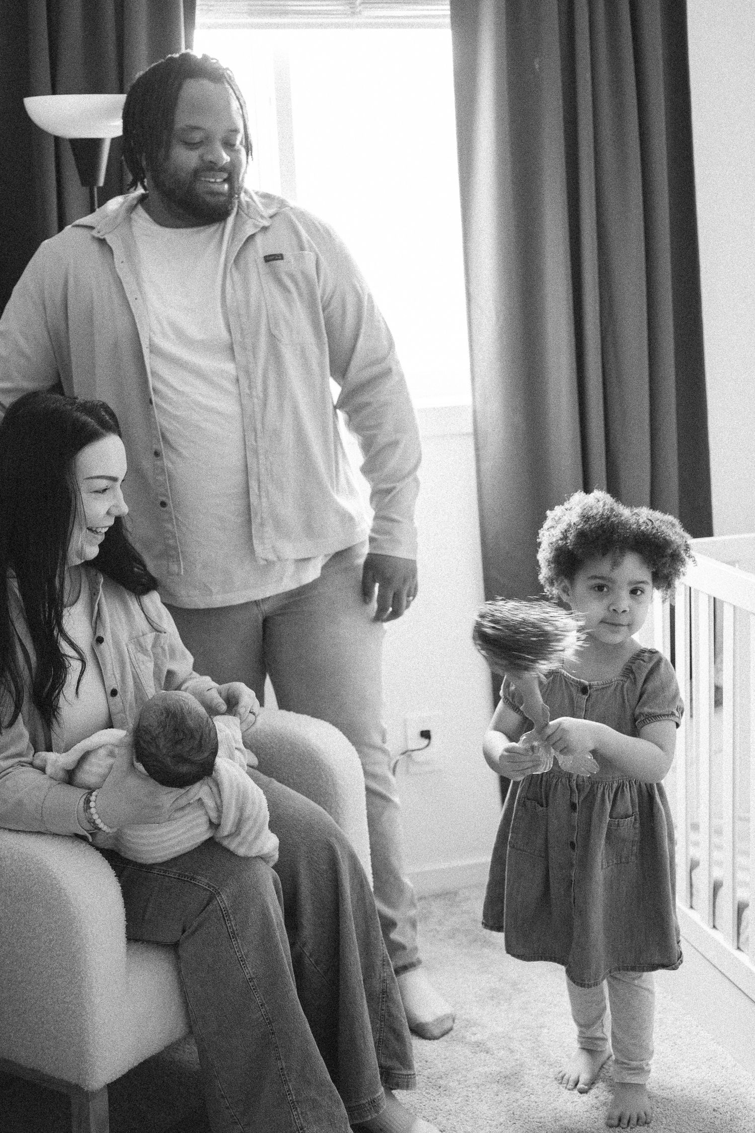 Black and white photo of a family with a mother holding a newborn, father standing behind, and toddler girl holding a toy.