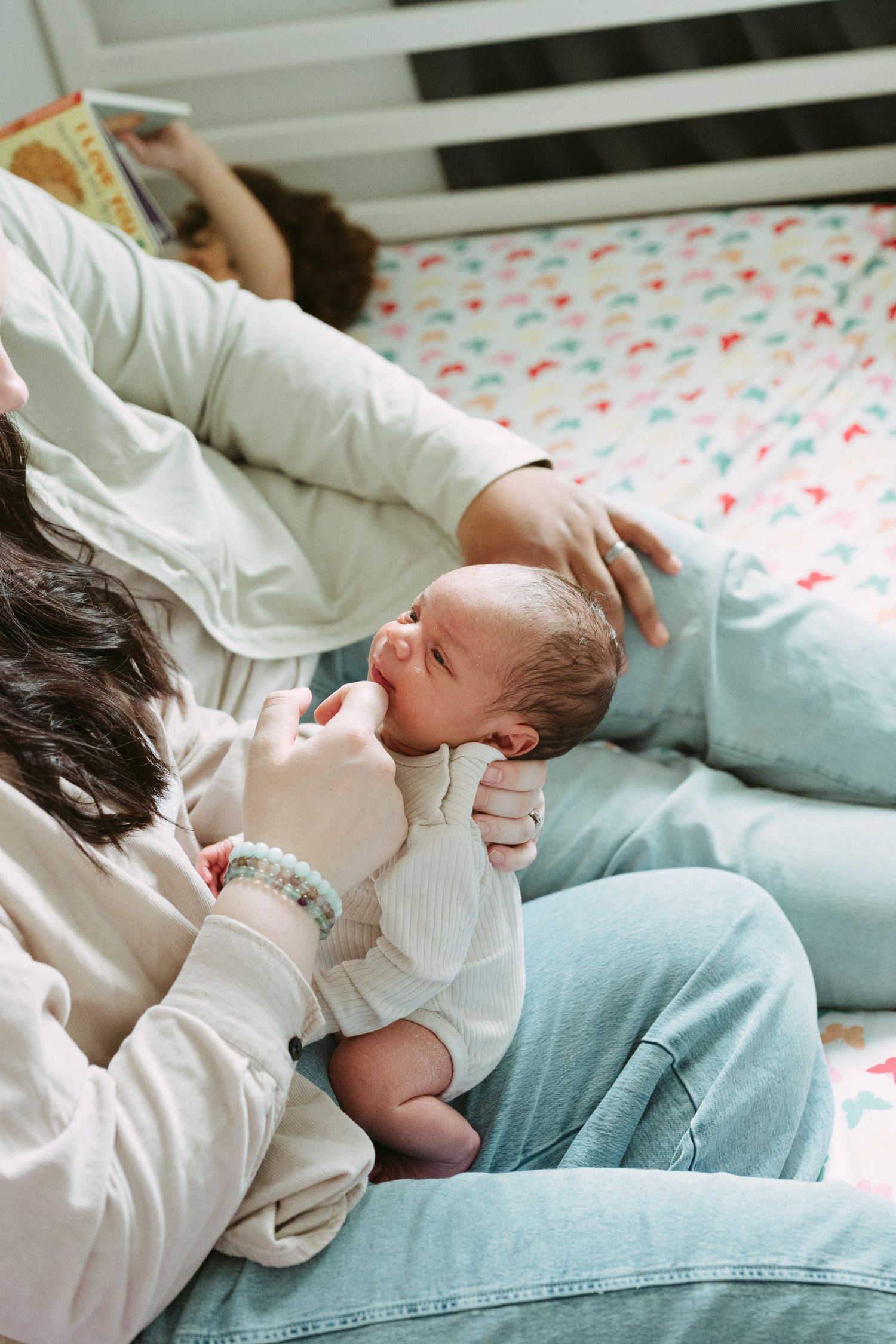 A mother holds her newborn baby on a couch while another child reads a book in the background.
