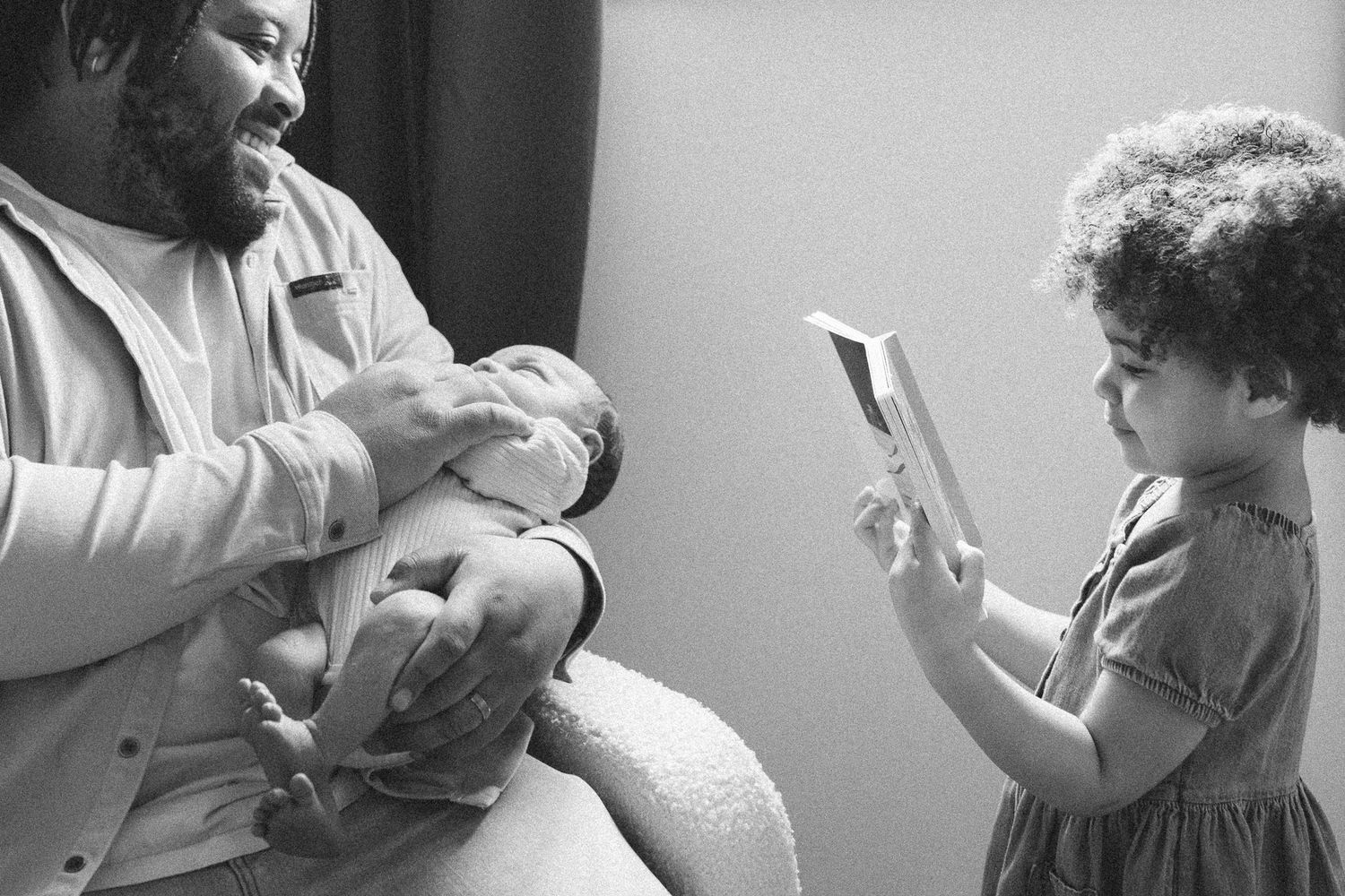 Black and white photo of father holding newborn while young girl shows him a book in the nursery.