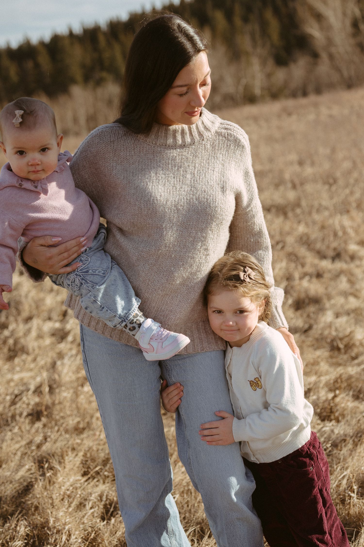 A parent wearing a beige sweater holds a baby in pink while another child in white hugs their leg in a grassy field.